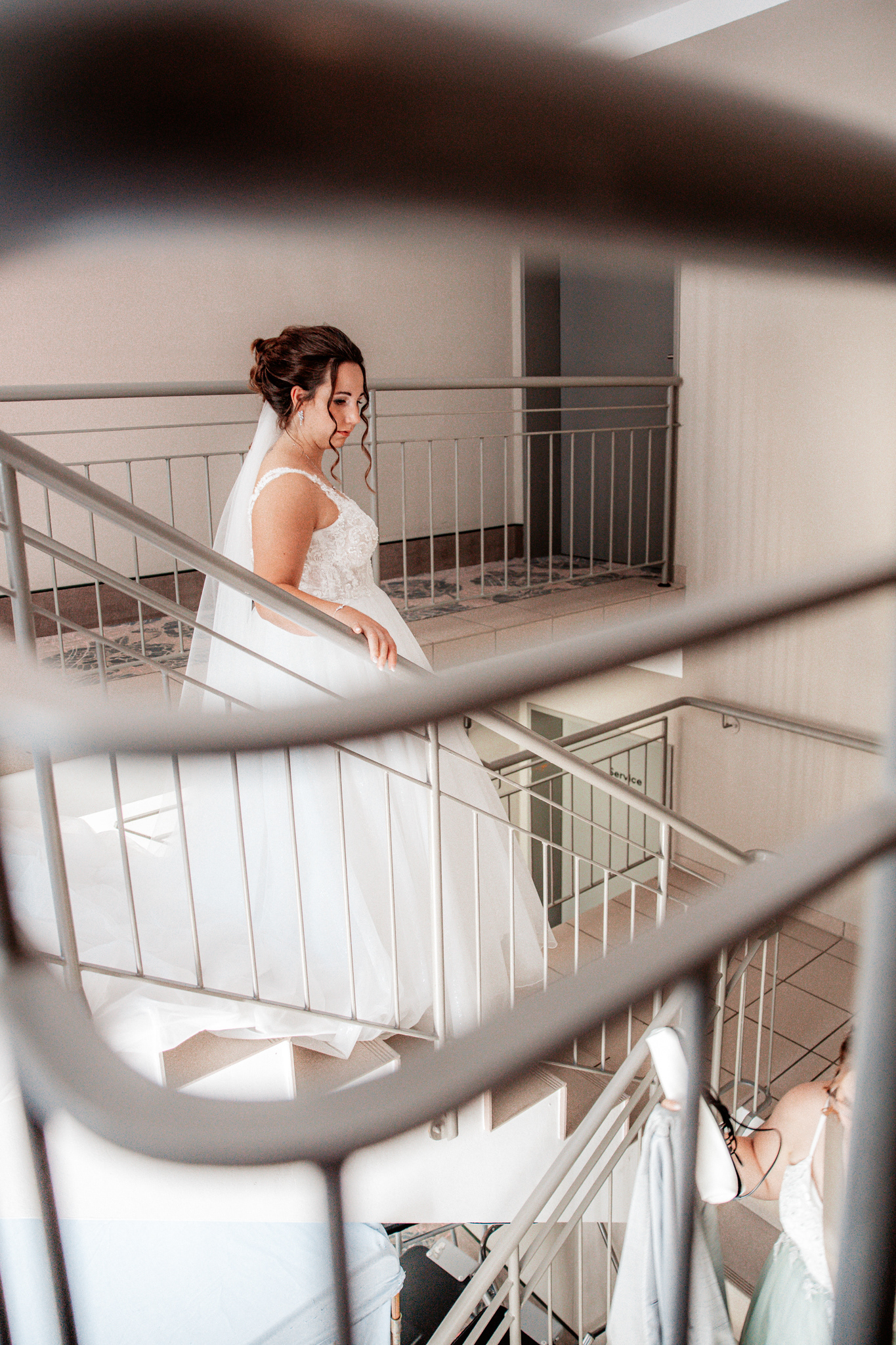 Braut Denise schreitet elegant die Treppe hinunter, ihr weißes Hochzeitskleid mit langer Schleppe fließt hinter ihr her. Die Szene ist durch ein Geländer eingefasst, was die Spannung und Vorfreude auf die Trauung verstärkt.