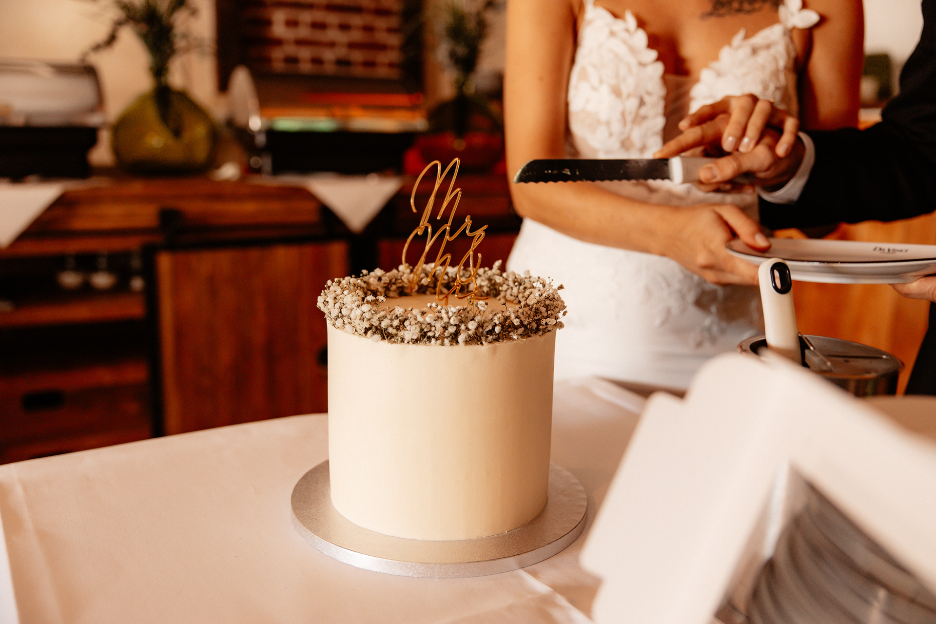 Das Brautpaar schneidet gemeinsam die Hochzeitstorte an – eine elegante, minimalistische Torte mit weißem Überzug und Blumenkranz-Deko.