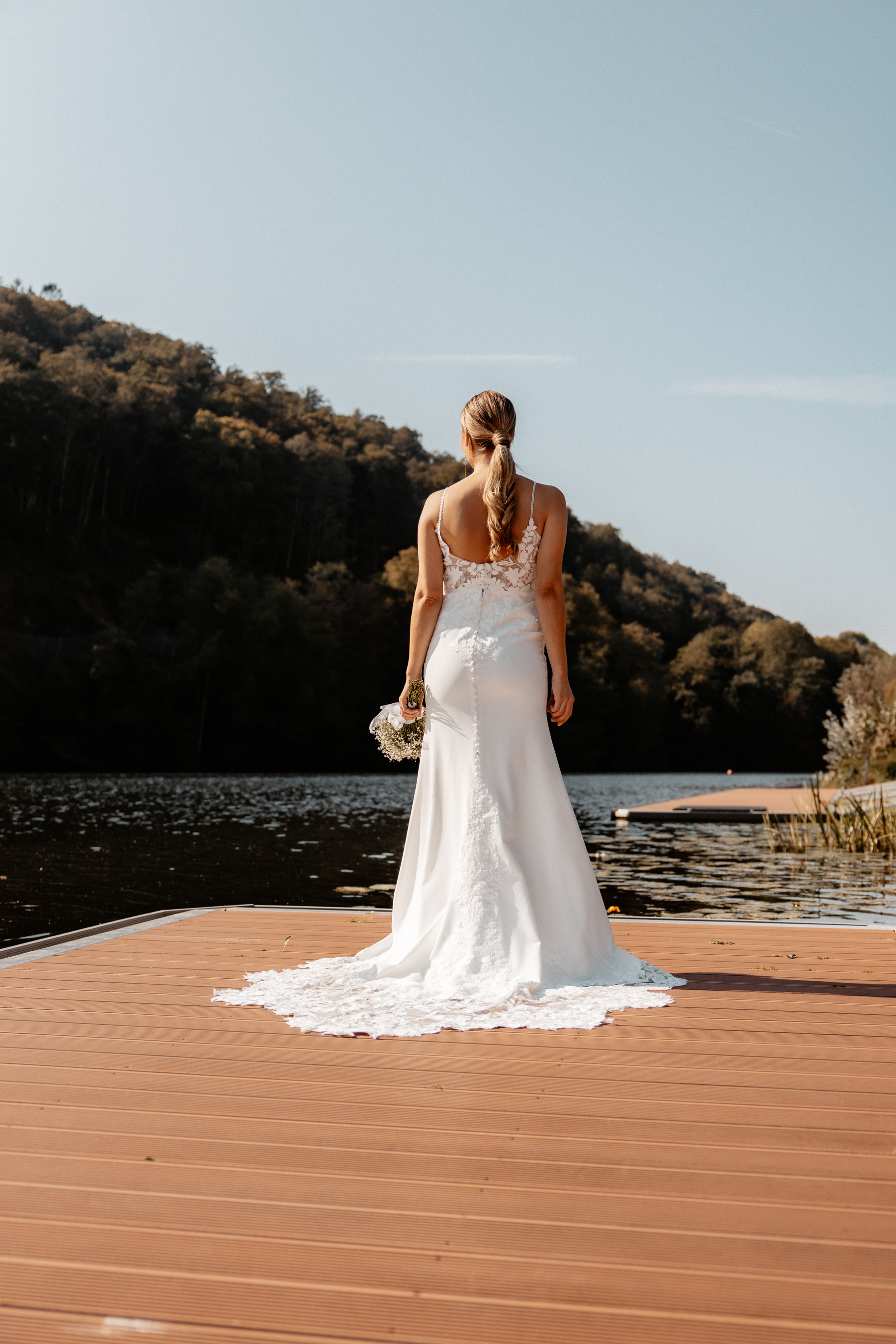 Romantische Rückenaufnahme der Braut auf einem Steg mit Blick auf den See, ihr langes Kleid fällt elegant über das Holz.