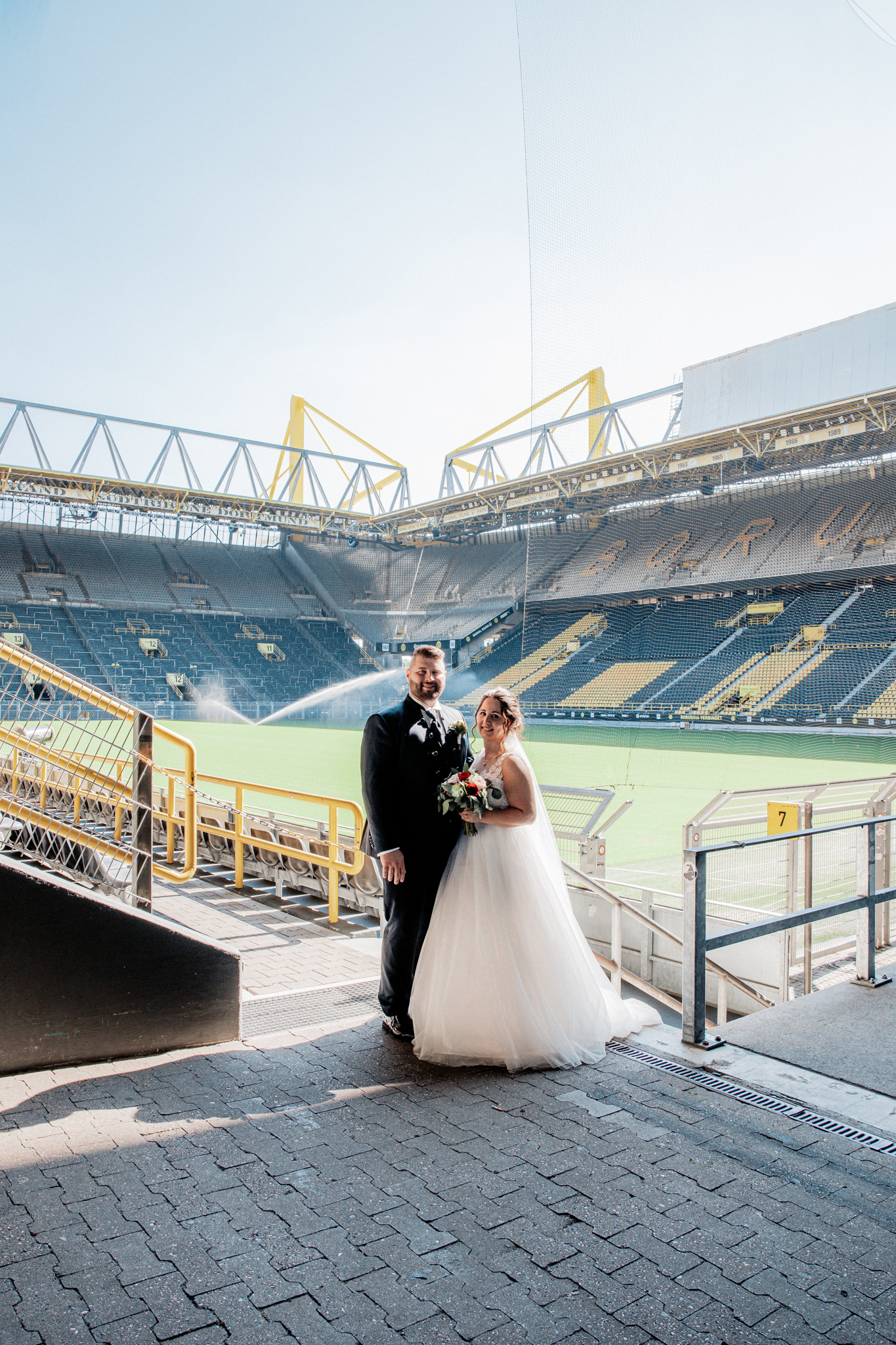 Hochzeitsfoto von Denise und Michael mit Blick auf das Stadioninnere des Signal Iduna Parks. Das Brautpaar steht auf der Treppe mit perfektem Blick auf das Spielfeld und die Tribünen der legendären BVB-Arena in Dortmund.