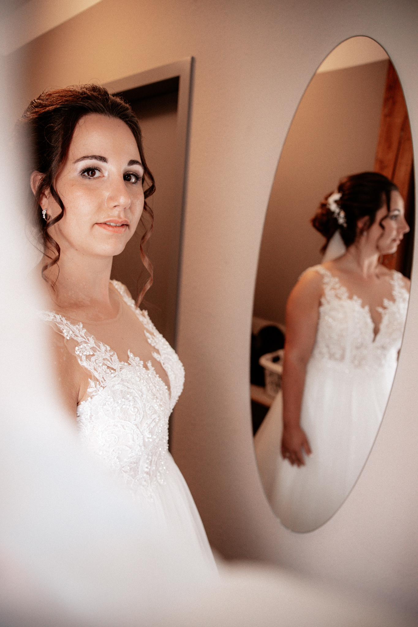 Braut Denise beim Getting Ready vor ihrem Hochzeitsspiegel. Ihr elegantes weißes Kleid mit filigraner Spitzenverzierung reflektiert sich im ovalen Spiegel, während sie mit sanftem Lächeln in die Kamera schaut. Eine romantische, intime Momentaufnahme vor der Hochzeit.