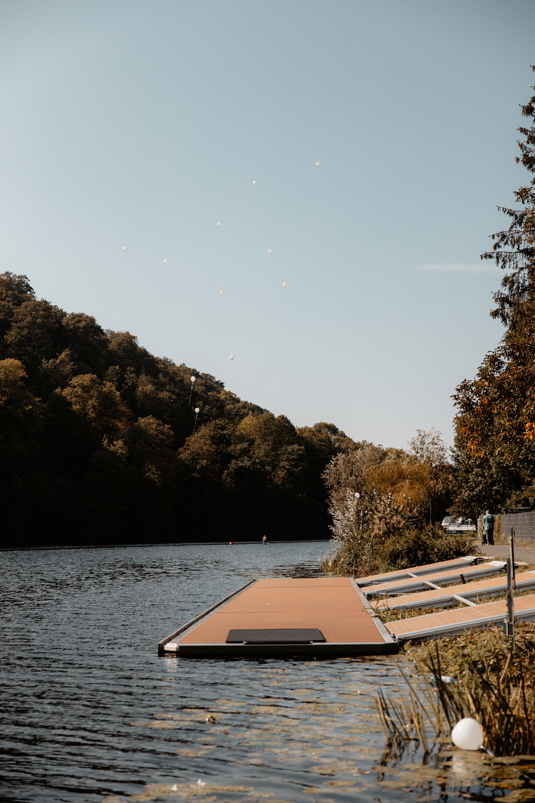 Weitwinkelaufnahme der Ballons, die langsam zwischen Himmel und Baumwipfeln verschwinden, während der Steg und das Wasser Ruhe ausstrahlen.