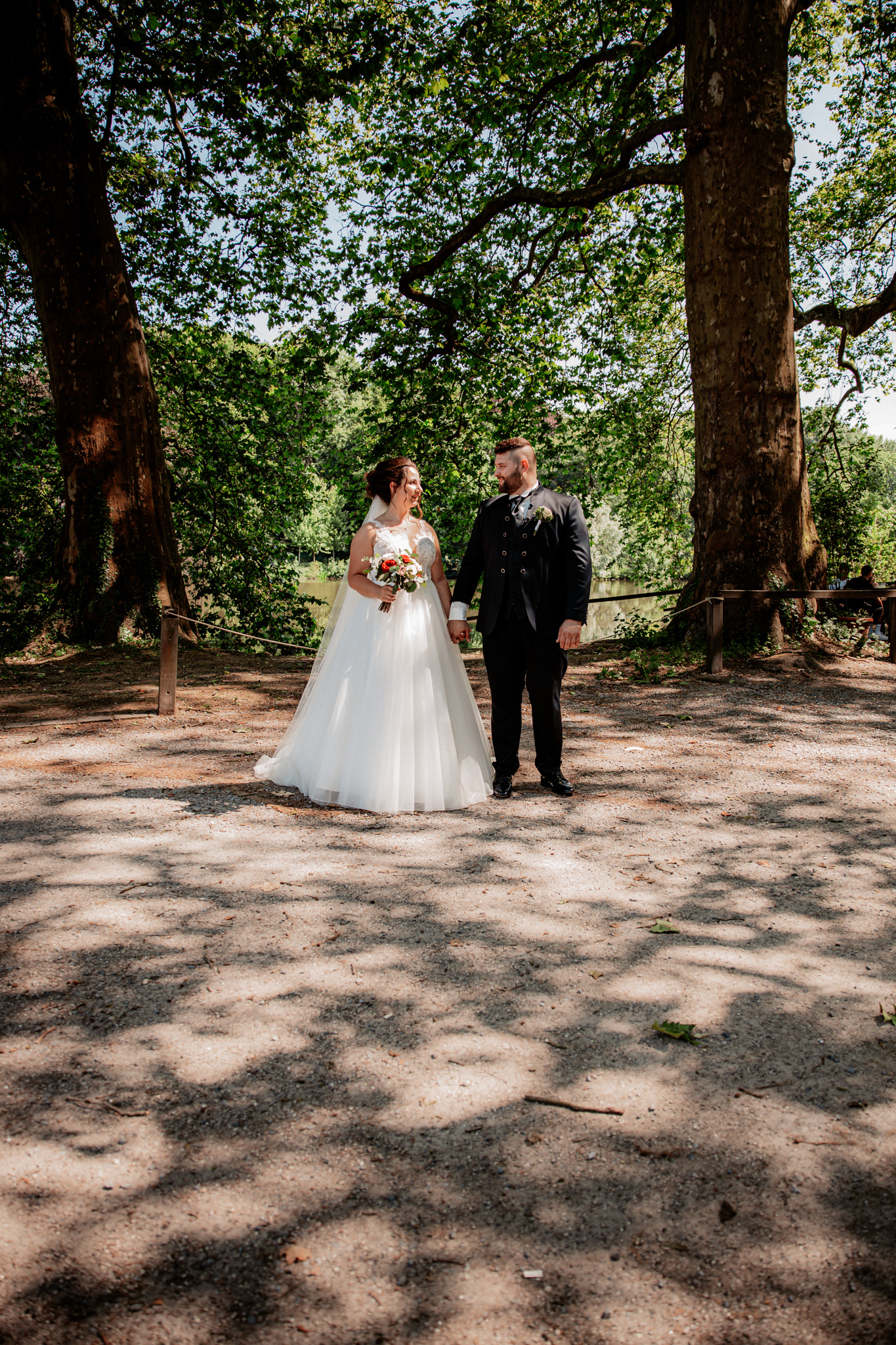 Denise und Michael stehen unter einem großen, schattenspendenden Baum in einem idyllischen Park in Dortmund. Die Sonne scheint durch das Laub, während sie sich verliebt ansehen und der Bräutigam die Hand der Braut hält.
