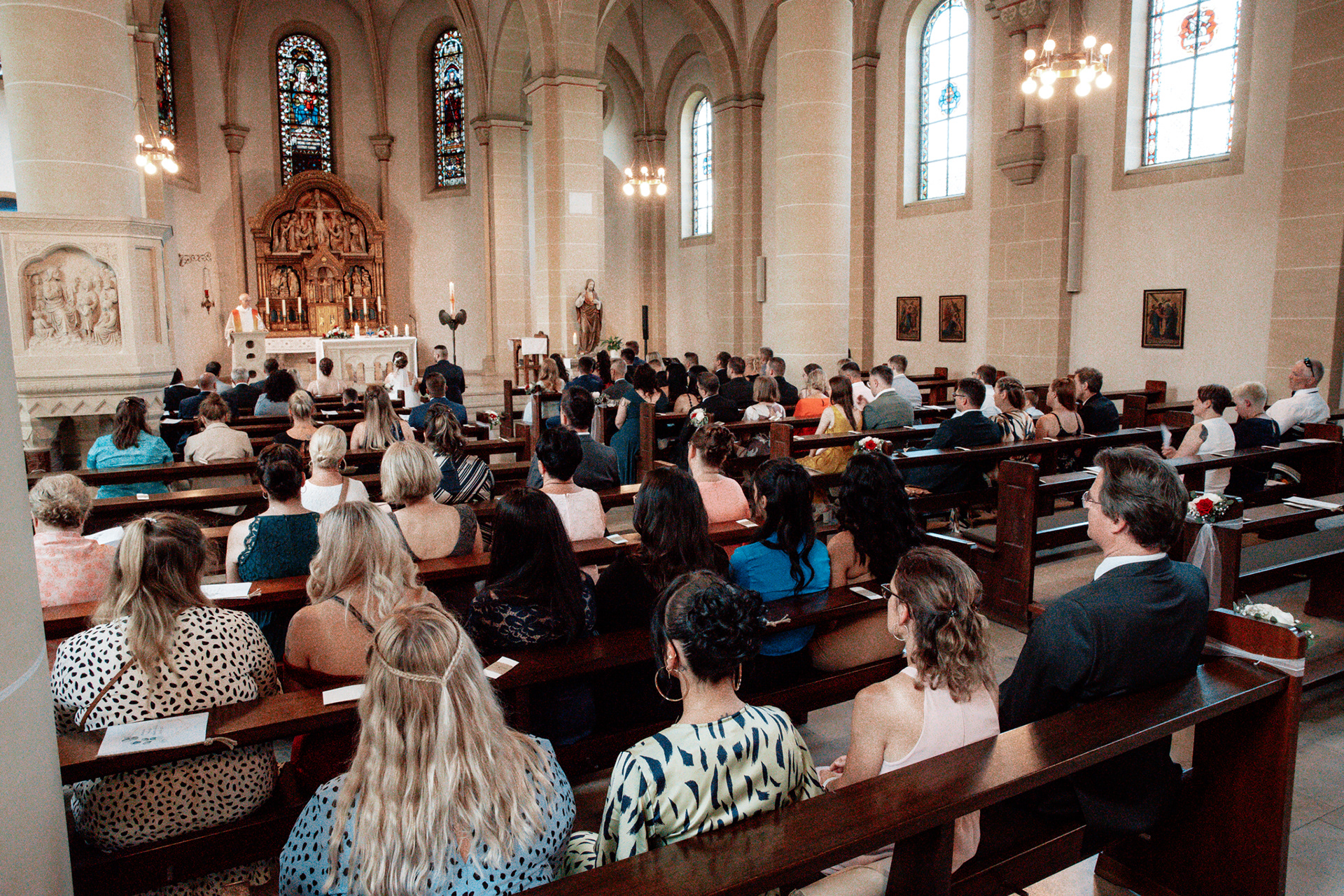 Die Hochzeitsgesellschaft von Denise und Michael sitzt in einer prachtvollen Kirche mit hohen Bögen und bunten Kirchenfenstern. Der Priester hält die Trauung vor einem kunstvollen Altar ab, während die Gäste aufmerksam zuhören.