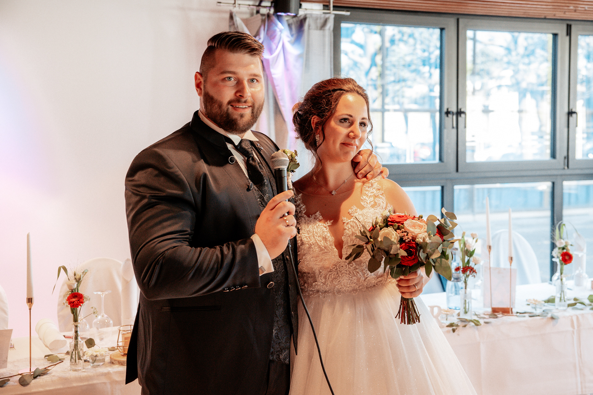 Feierlicher Moment während der Hochzeitsrede von Denise und Michael. Der Bräutigam hält ein Mikrofon und bedankt sich bei den Gästen, während die Braut mit ihrem Blumenstrauß glücklich an seiner Seite steht. Die Location ist stilvoll dekoriert mit eleganten Hochzeitsdetails.