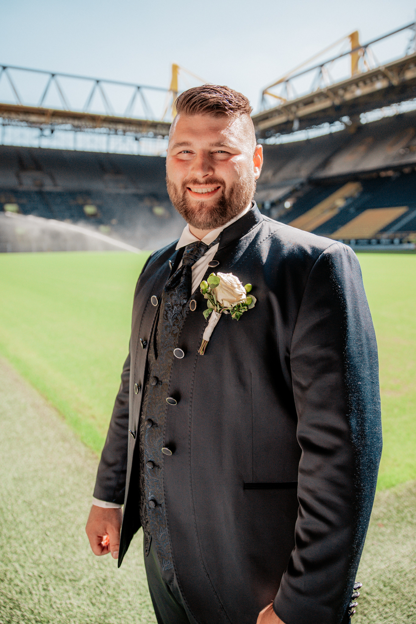 Strahlender Bräutigam im Signal Iduna Park Dortmund. Michael posiert stolz in seinem eleganten Hochzeitsanzug auf dem grünen Spielfeld des legendären Fußballstadions von Borussia Dortmund, während im Hintergrund die Sprinkleranlage das Spielfeld bewässert.