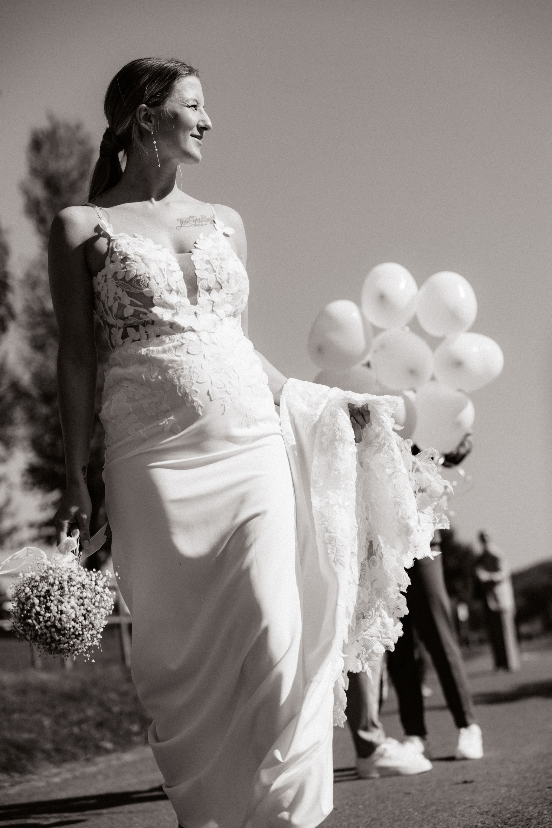 Schwarz-Weiß-Fotografie der Braut Justine, die mit einem strahlenden Lächeln durch den Sonnenschein schreitet, in der Hand ihren Brautstrauß und das Kleid leicht angehoben.