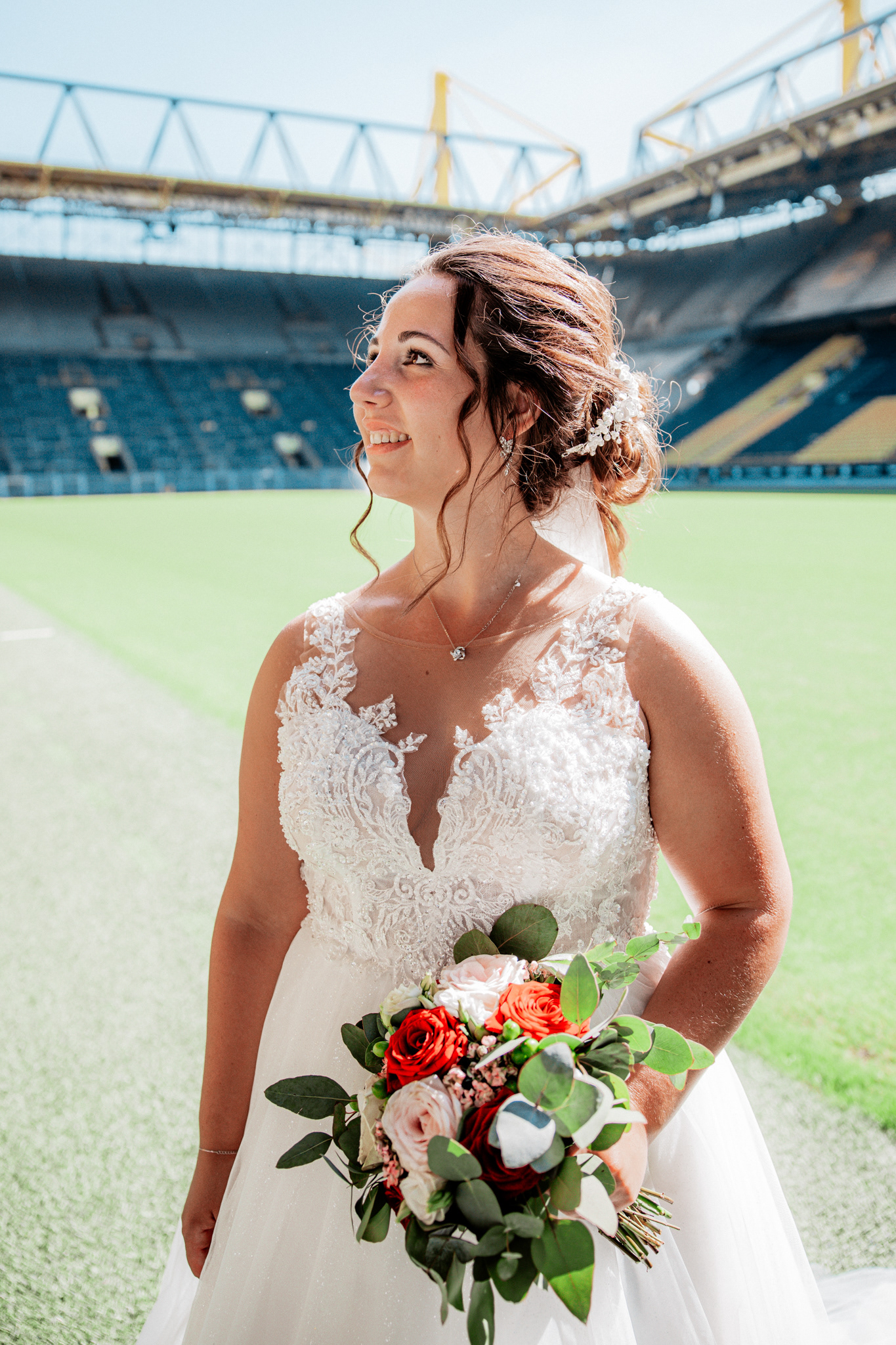 Romantische Hochzeitsaufnahme von Denise und Michael im Dortmunder Signal Iduna Park. Die Braut hält ihren stilvollen Brautstrauß, während sie glücklich in die Ferne blickt, mit dem beeindruckenden Stadionhintergrund der BVB-Arena.
