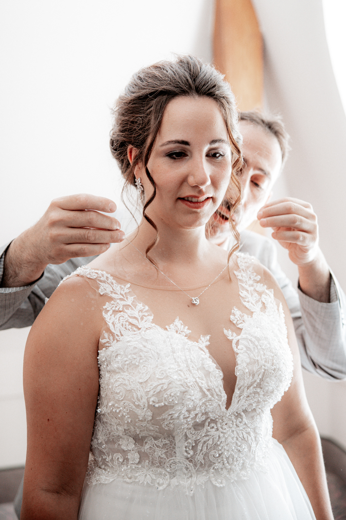 Braut Denise erhält eine zarte Silberkette als Geschenk während des Getting Ready. Der Vater hilft ihr, das Schmuckstück um den Hals zu legen. Sie trägt ein elegantes Hochzeitskleid mit filigraner Spitze und strahlt eine Mischung aus Freude und Aufregung aus.