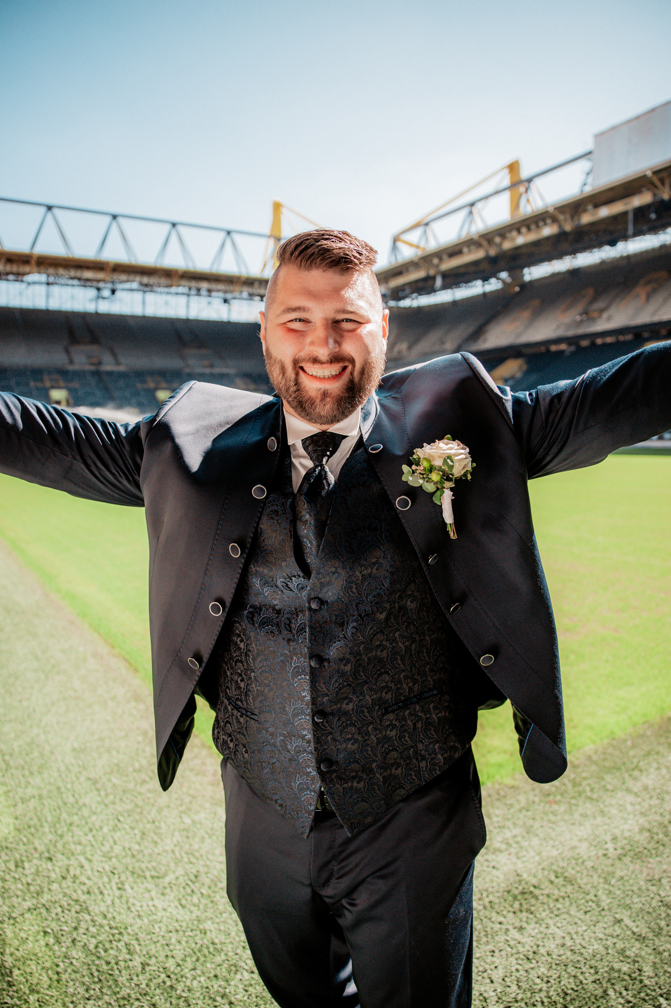 Freudiger Moment des Bräutigams im BVB-Stadion in Dortmund. Michael breitet voller Begeisterung die Arme aus, während er im festlichen Hochzeitsoutfit auf dem Spielfeld des Signal Iduna Parks steht, bereit für einen neuen Lebensabschnitt.