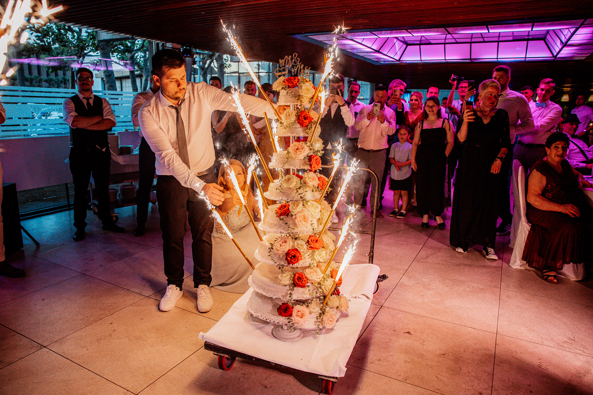 Spektakuläre, mehrstöckige Hochzeitstorte von Denise und Michael in der Europahalle Castrop-Rauxel. Die imposante Torte besteht aus sieben kunstvoll verzierten Stufen, dekoriert mit weißen und roten Rosen. Mehrere brennende Wunderkerzen sorgen für eine feierliche Atmosphäre, während der Bräutigam nach der Torte greift und Gäste den Moment mit ihren Handys festhalten.
