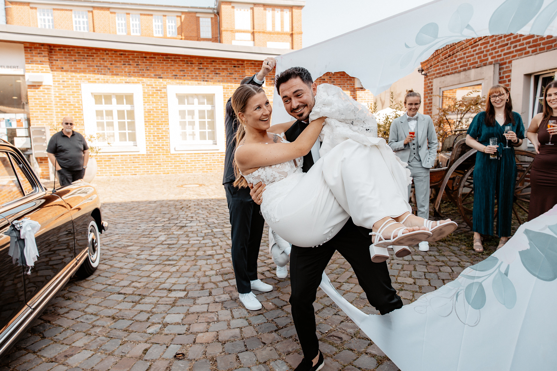 Der Bräutigam trägt seine strahlende Braut durch das zuvor ausgeschnittene Herz im Hochzeitstuch, während die Gäste lachend und mit Getränken in der Hand das Paar feiern.