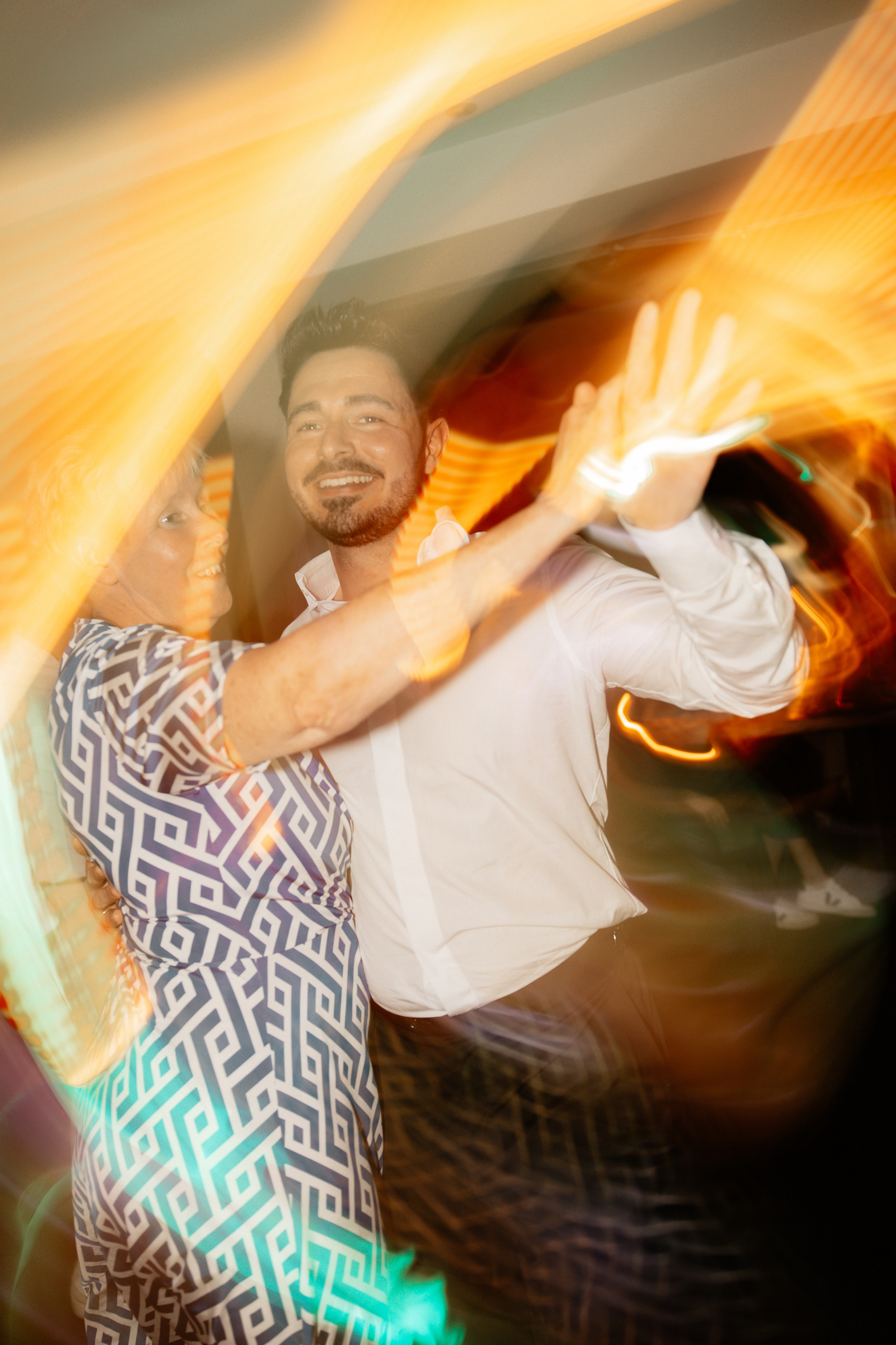 Der Bräutigam tanzt mit einer seiner Mutter, beide lachen herzlich, während farbige Lichtstreifen die Bewegung auf der Tanzfläche einfangen.