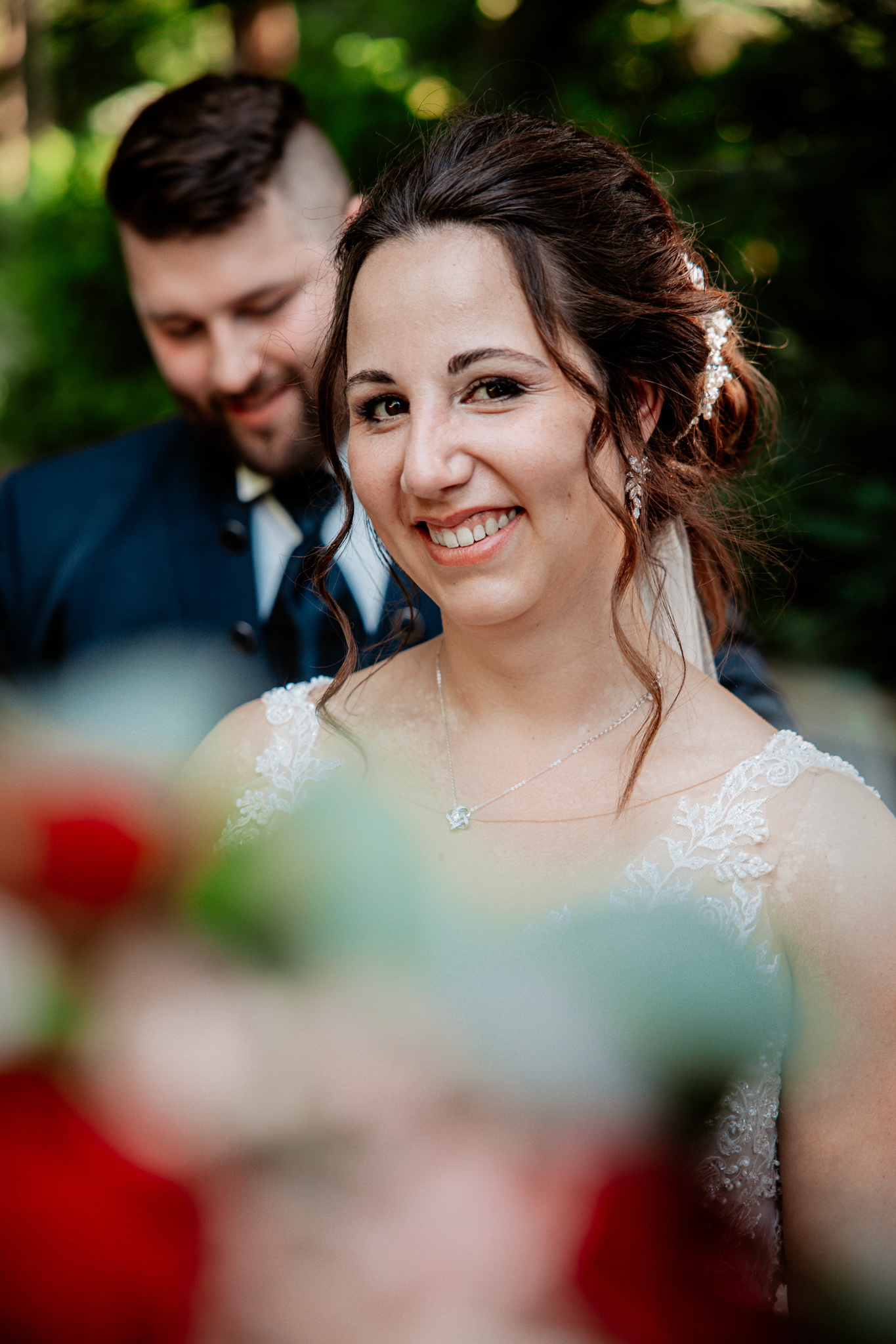 Die Braut strahlt vor Glück mit einem unscharfen Blumenstrauß im Vordergrund, während der Bräutigam im Hintergrund lächelt.