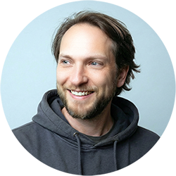 Smiling man with short brown hair and a trimmed beard wearing a dark hoodie, looking slightly to the side against a light background