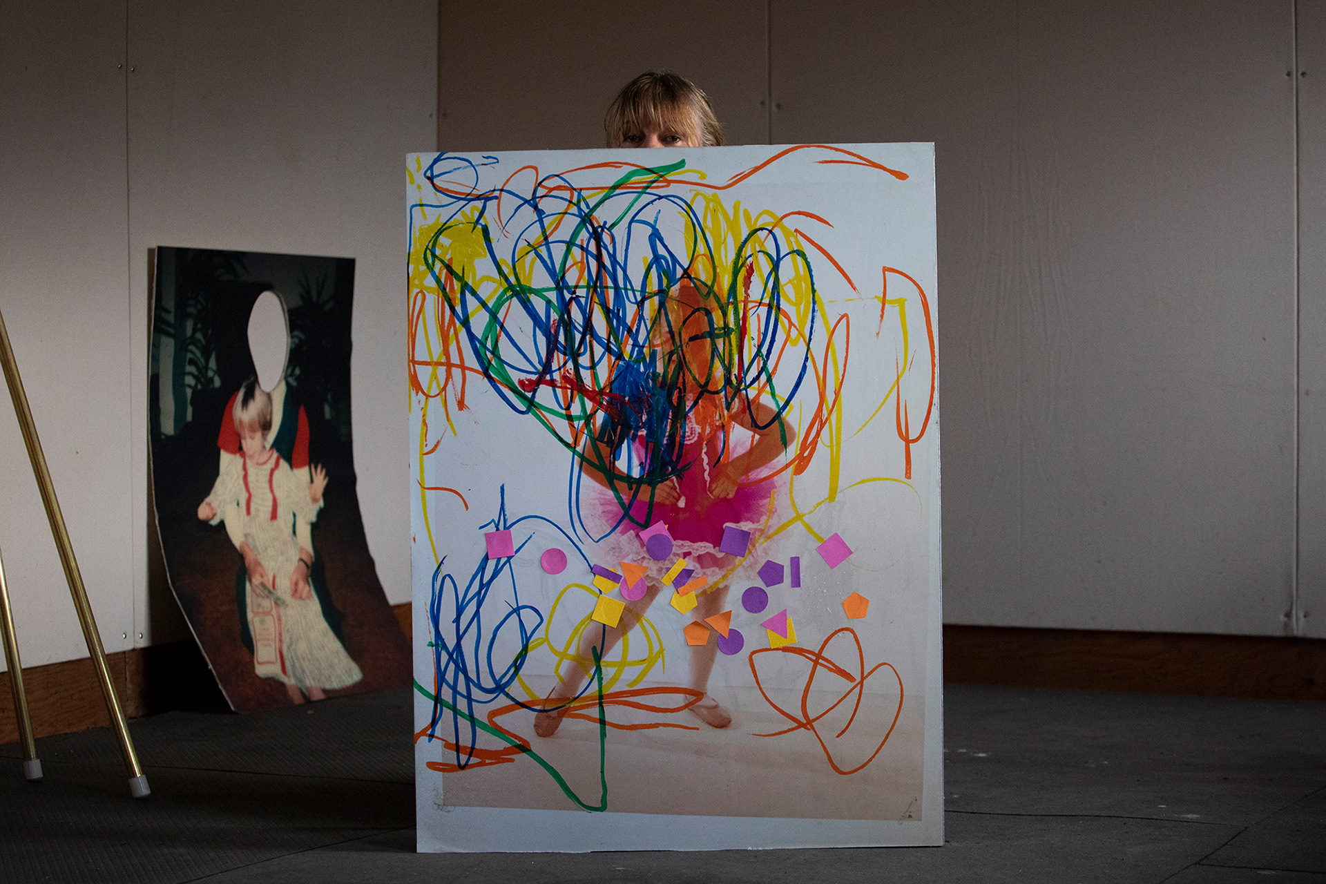 Renee Couture, a multi-media artist from Glide, Ore., peaks over a piece she created during her residency at Pine Meadow Ranch in Sisters, Ore., on Nov. 3, 2021. The piece showcases a photo of Couture as a young child in a pink tutu that was later covered by paint, shapes and other markings by her daughter. Couture created the piece with her daughter—who is a same age as she was when the photo was taken—to explore what it means to be young and the differences and similarities between her and her child.