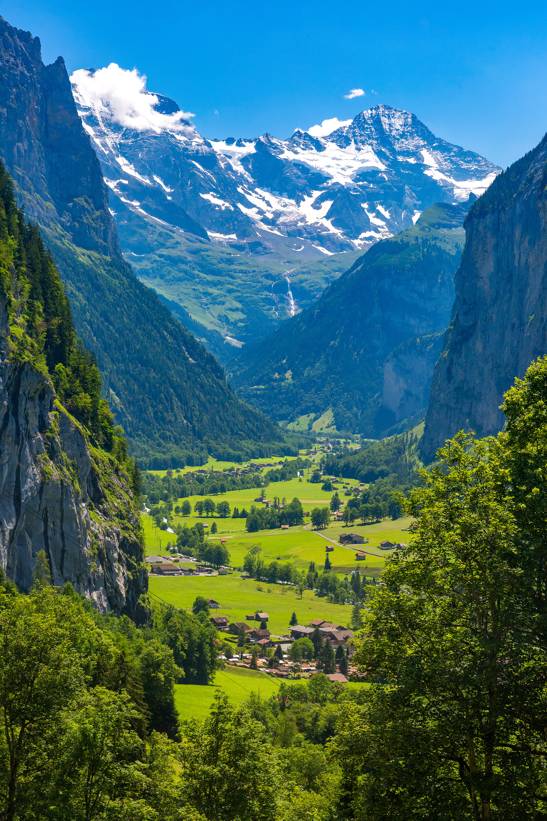 A breathtaking view of a Swiss valley with lush green meadows and chalets, flanked by steep cliffs and majestic snow-capped mountains, embodying the origin and inspiration behind Pan_Inc’s HandPans.