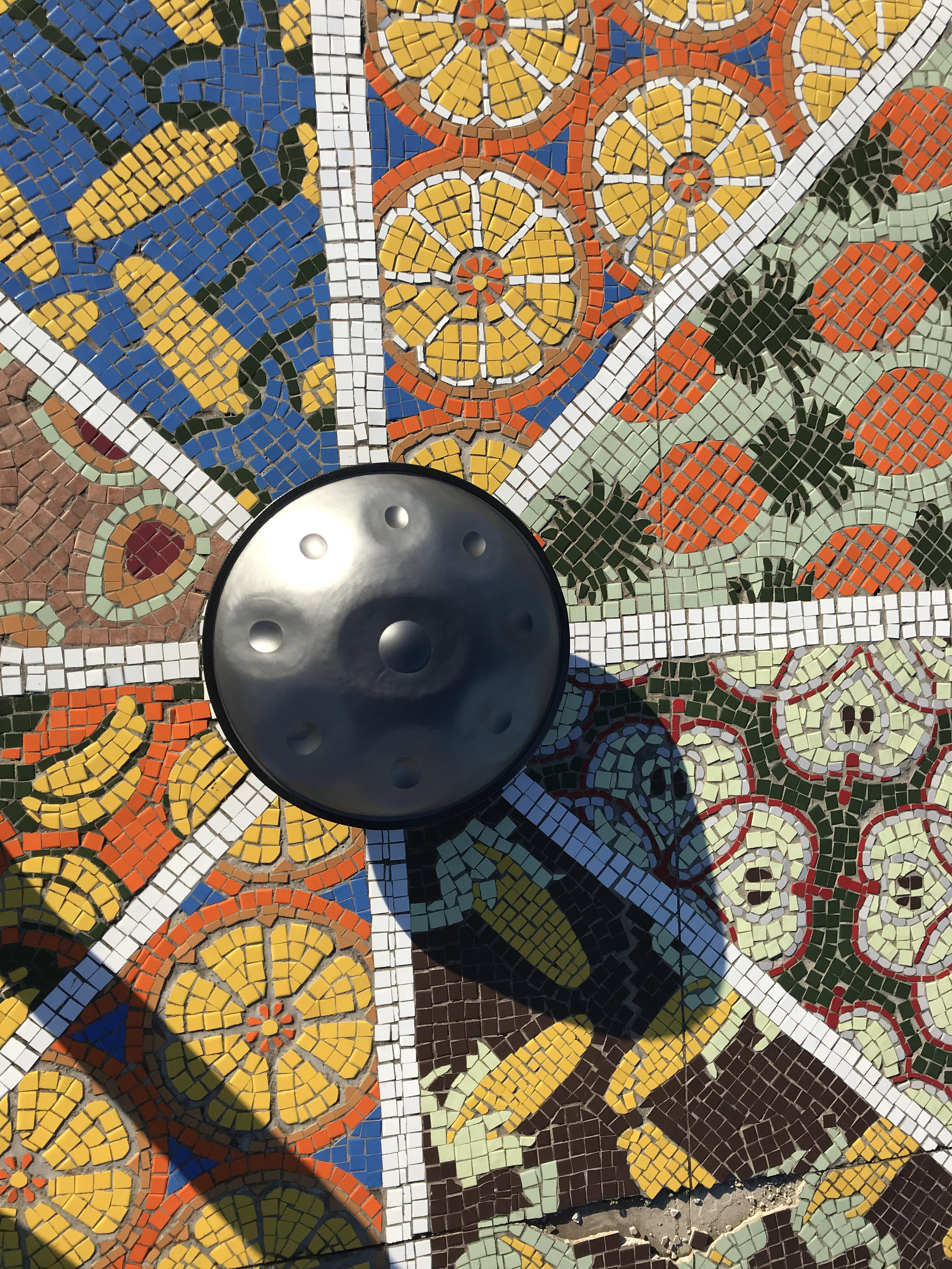 The Pan_Inc Authentic Series HandPan rests atop an elaborate mosaic, its sleek steel surface contrasting with the vibrant tiles, symbolizing a fusion of traditional art and modern music.