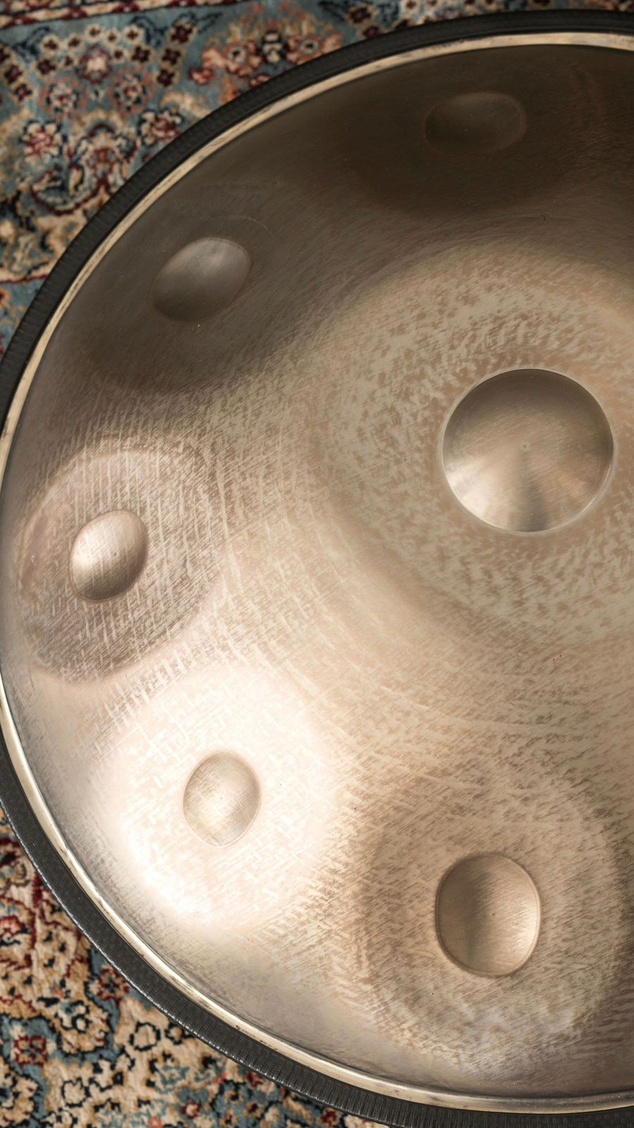 A close-up of a Pan_Inc Stainless Series HandPan, displaying its lustrous metal surface with crafted indentations, all set against the backdrop of an ornate rug.