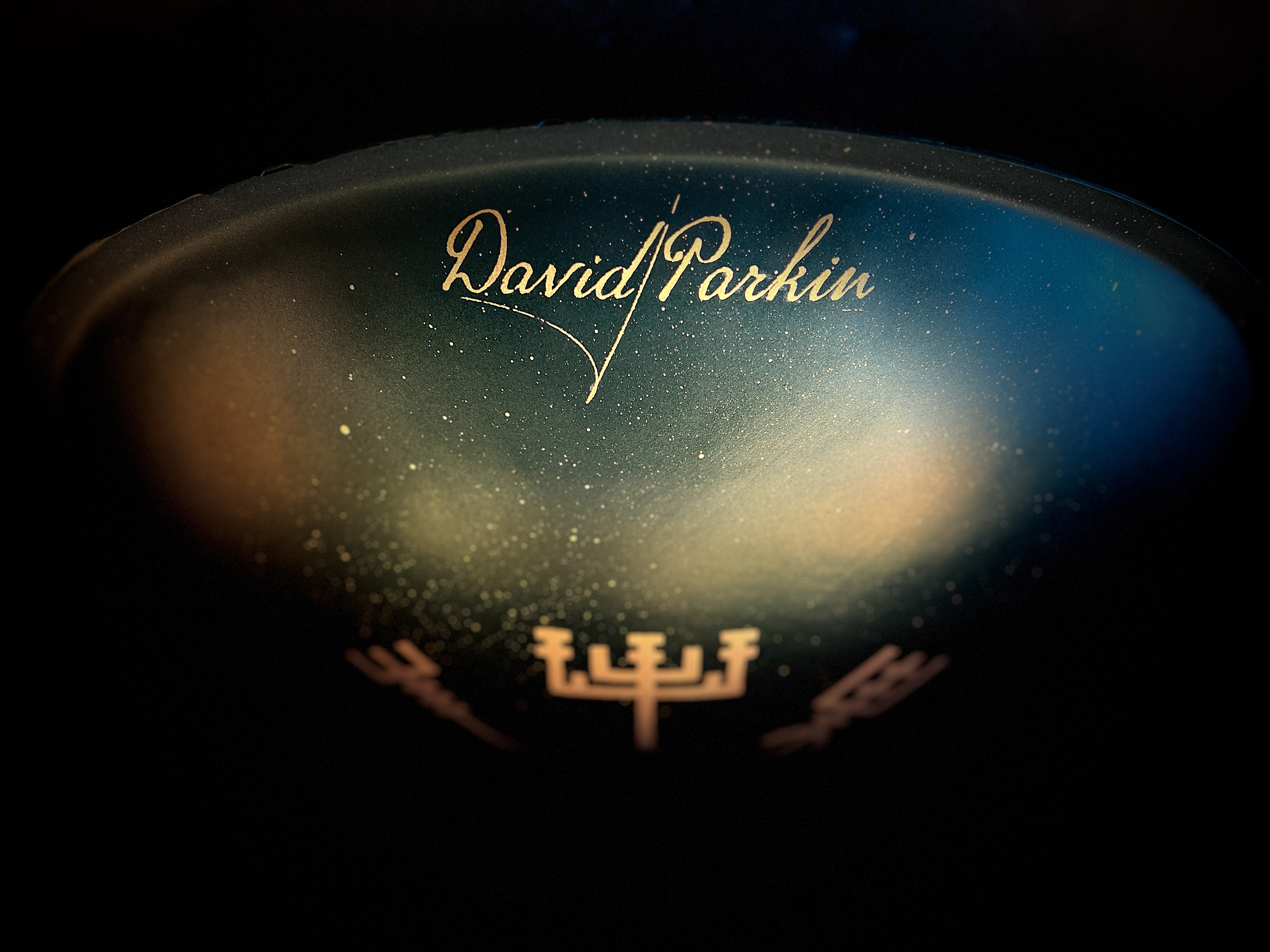 The gleaming surface of a Pan_Inc Custom Series HandPan, featuring the golden signature of David Parkin, set against a starry backdrop that evokes the celestial inspiration behind the instrument’s creation.