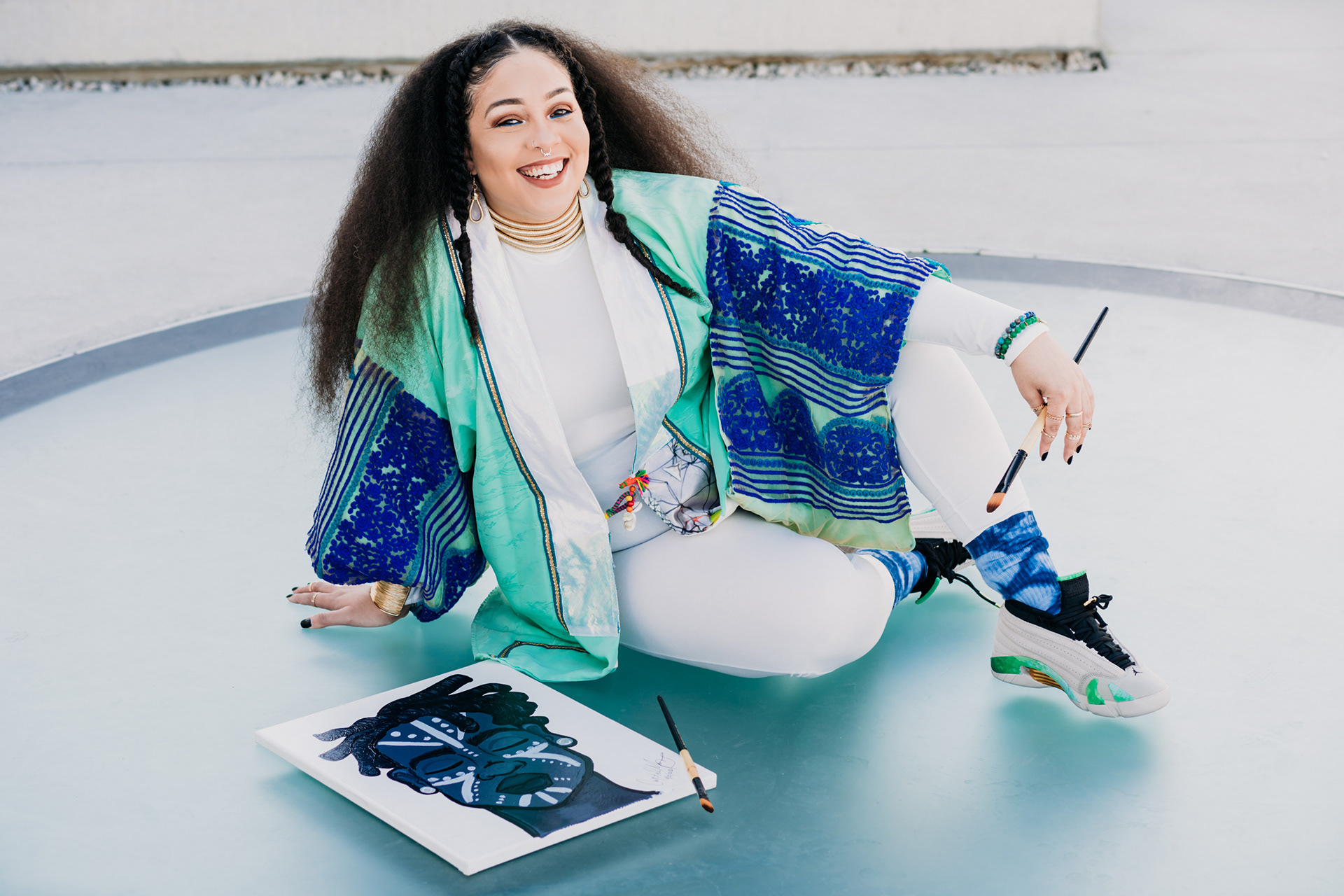 Photo of Crystal Manning a Houston based Artist and Illustrator wearing a tiffany blue and royal blue Kimono with gold jewelry and Air Jordan Sneaker. Crystal is sitting on the ground, posing next to a black and white painting of a man with locs and tribal face painting. Crystal is smiling happily and holding paint brushes.