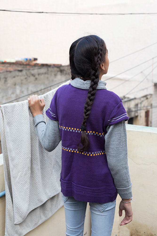 Riya stood on her roof with her back to the camera. She is holding onto a washing line and has a long plait down the right side of her back.