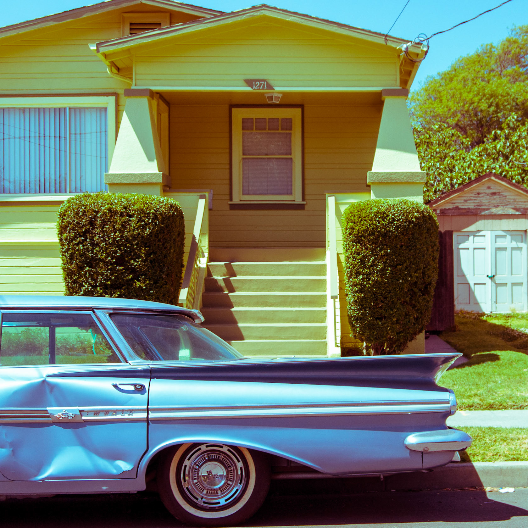 Dented blue Chevy Impala parked in front of a fabulous green house with bushes. Oakland CA. 2008.