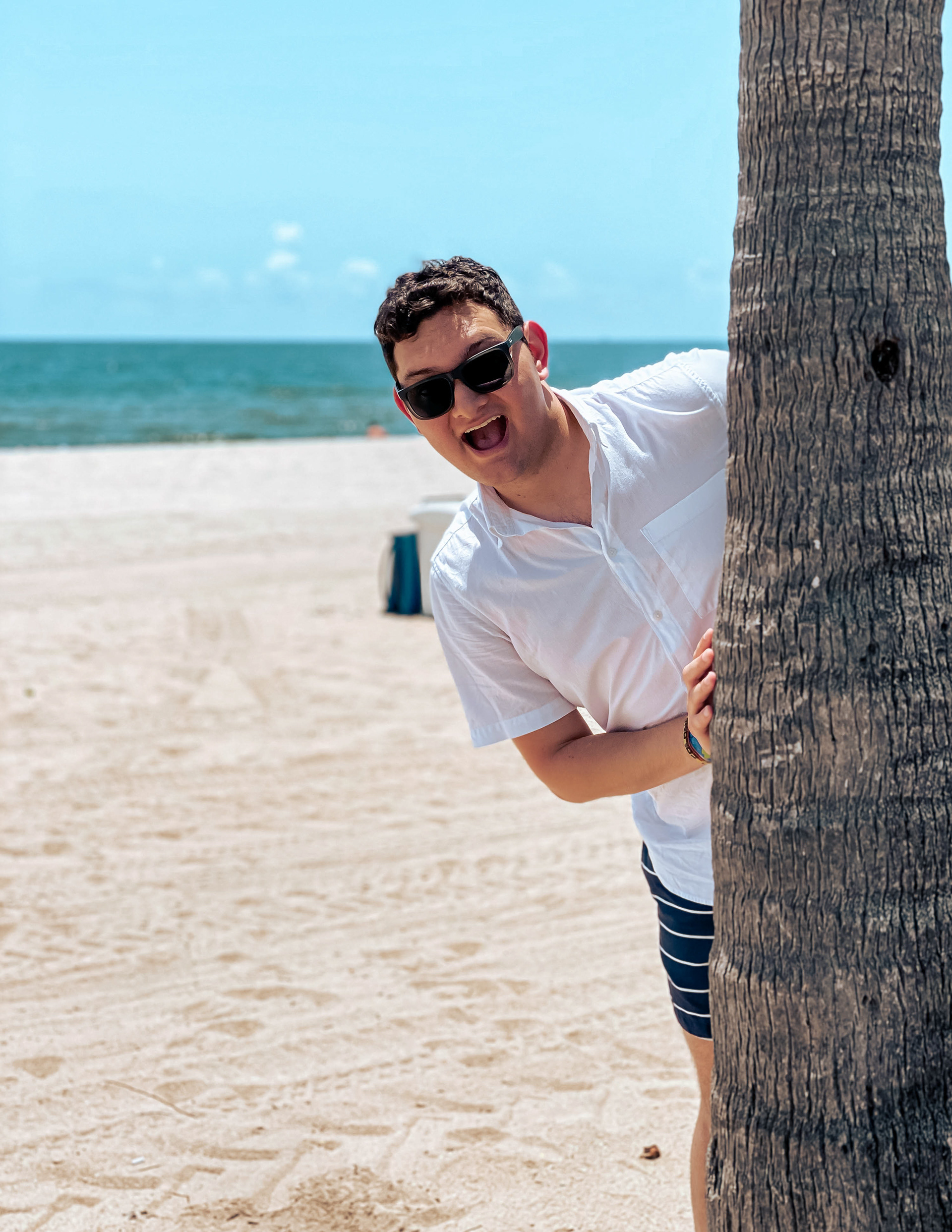 Photo of Milo Estevez smiling with sunglasses at the beach. Half of Milo's body is hiding behind a palm tree.