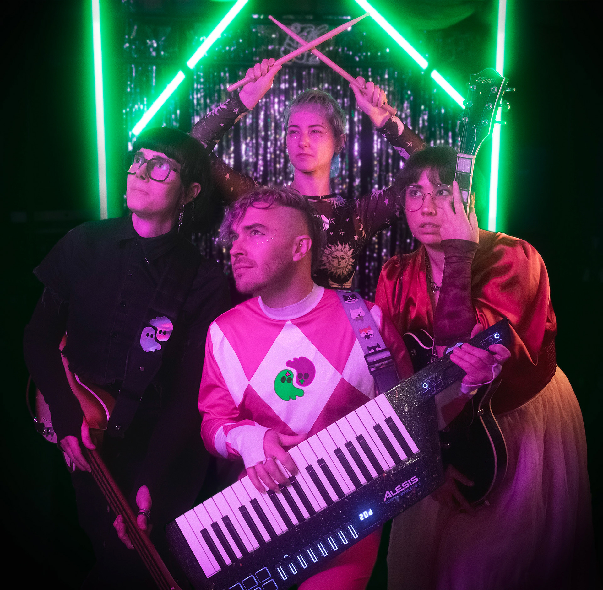 4 curiously dressed musicians stare off-screen to the top left. They are bathed in a soft pink light, with a vibrant glowing green stage in the background.