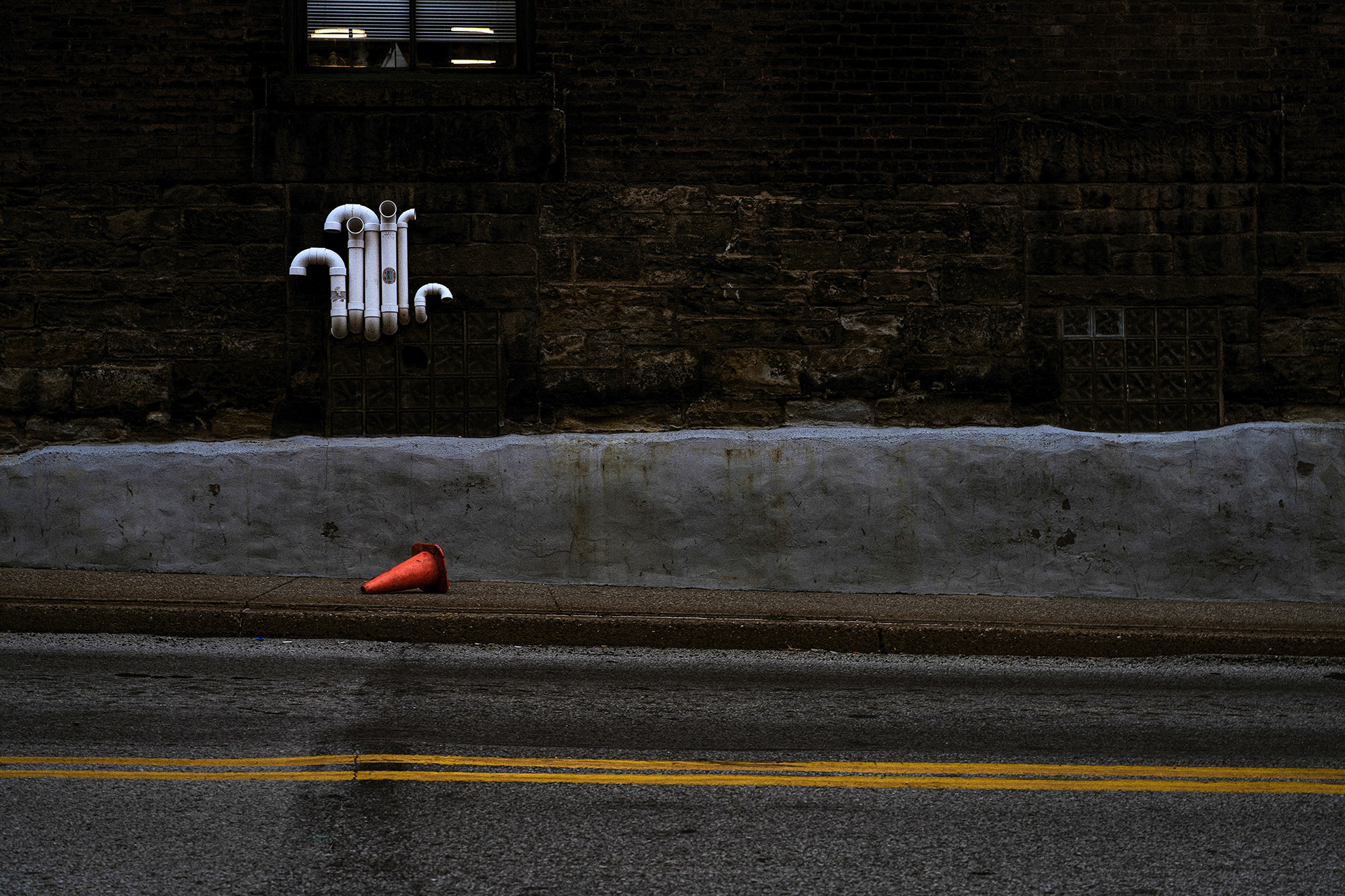 Fallen orange street cone by road with yellow lines in early morning by Bucchianeri.