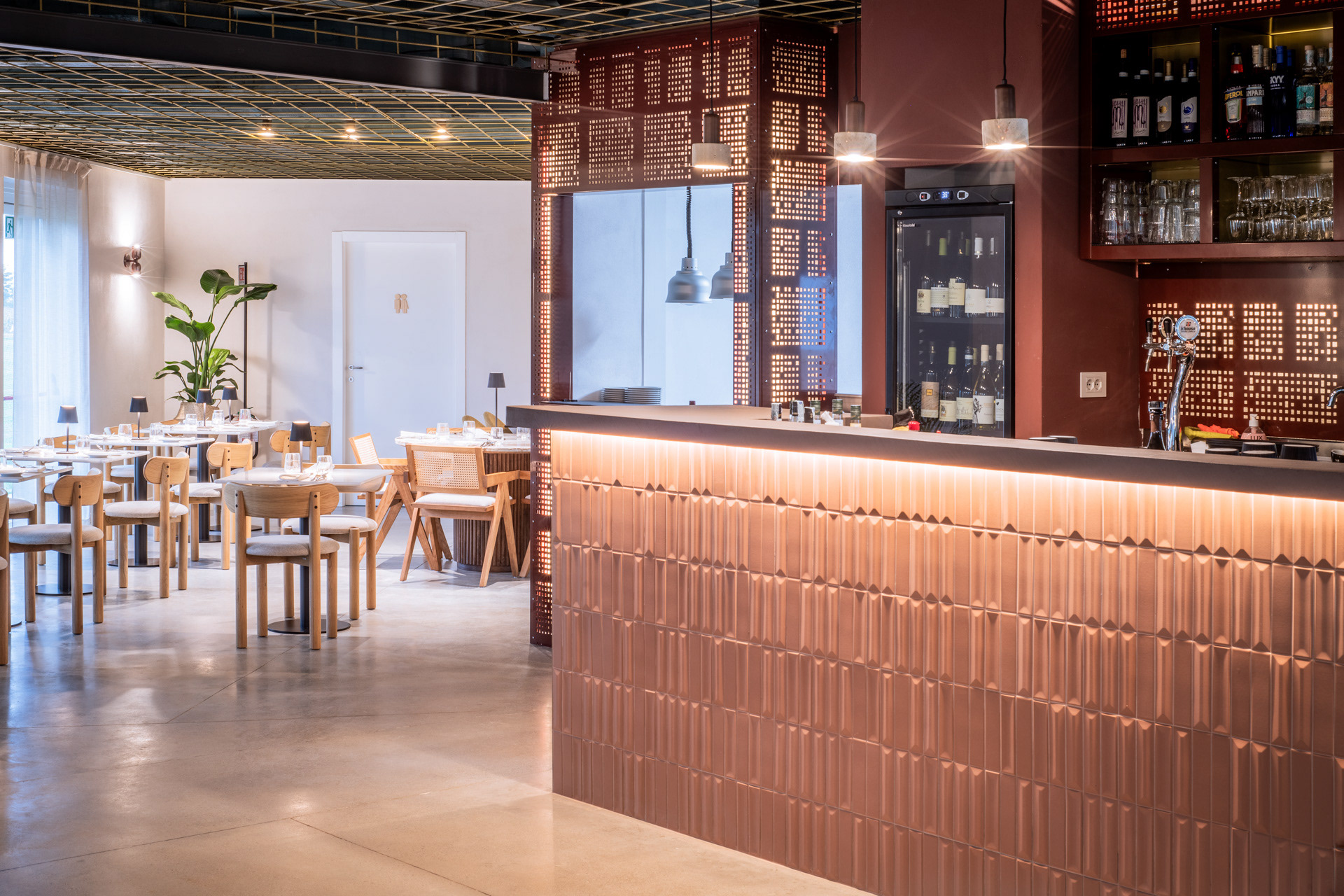 Interno del ristorante Archipelago con bancone bar rivestito con piastrelle 3d color terracotta, mentre il retro bar con lamiera con texture tridimensionale color porpora e illuminazione LED integrata. Griglia metallica forata retroilluminata che separa l’area bar dalla sala. Soffitto con griglia ottonata a contrasto con il blu scuro. Arredamento in legno naturale, sedie con intreccio in paglia di Vienna e illuminazione d’atmosfera. Tavoli sempre in marmo bianco venato. L'illuminazione è calda e soffusa.