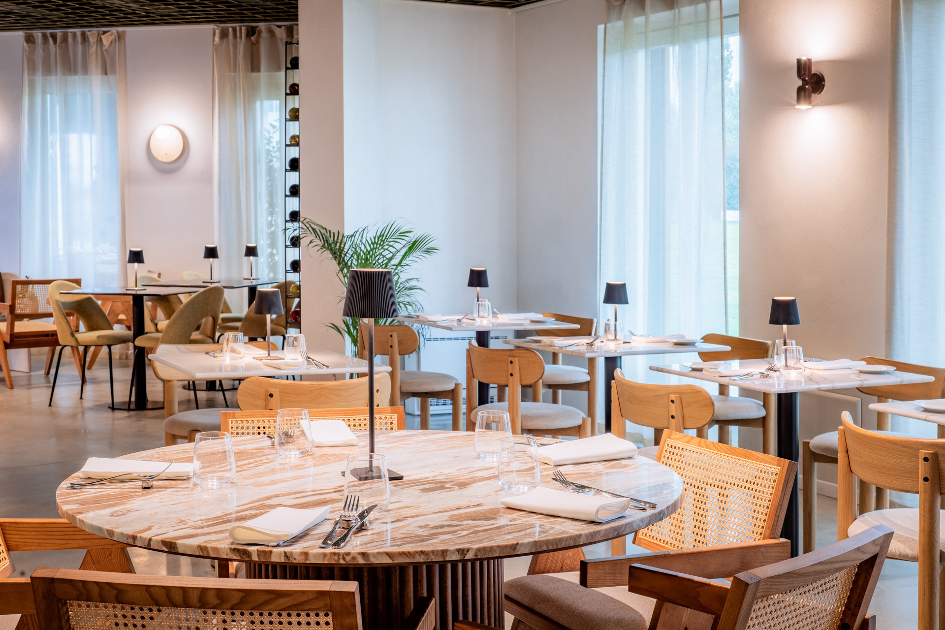 Interno del ristorante Archipelago con griglia metallica forata retroilluminata sulla sinistra, creando un effetto luminoso d'atmosfera. Il soffitto presenta una struttura a griglia ottonata che contrasta con il blu intenso dello sfondo. Tavoli in marmo, sedie in legno con intreccio in paglia di Vienna e luci soffuse completano il design elegante e raffinato.