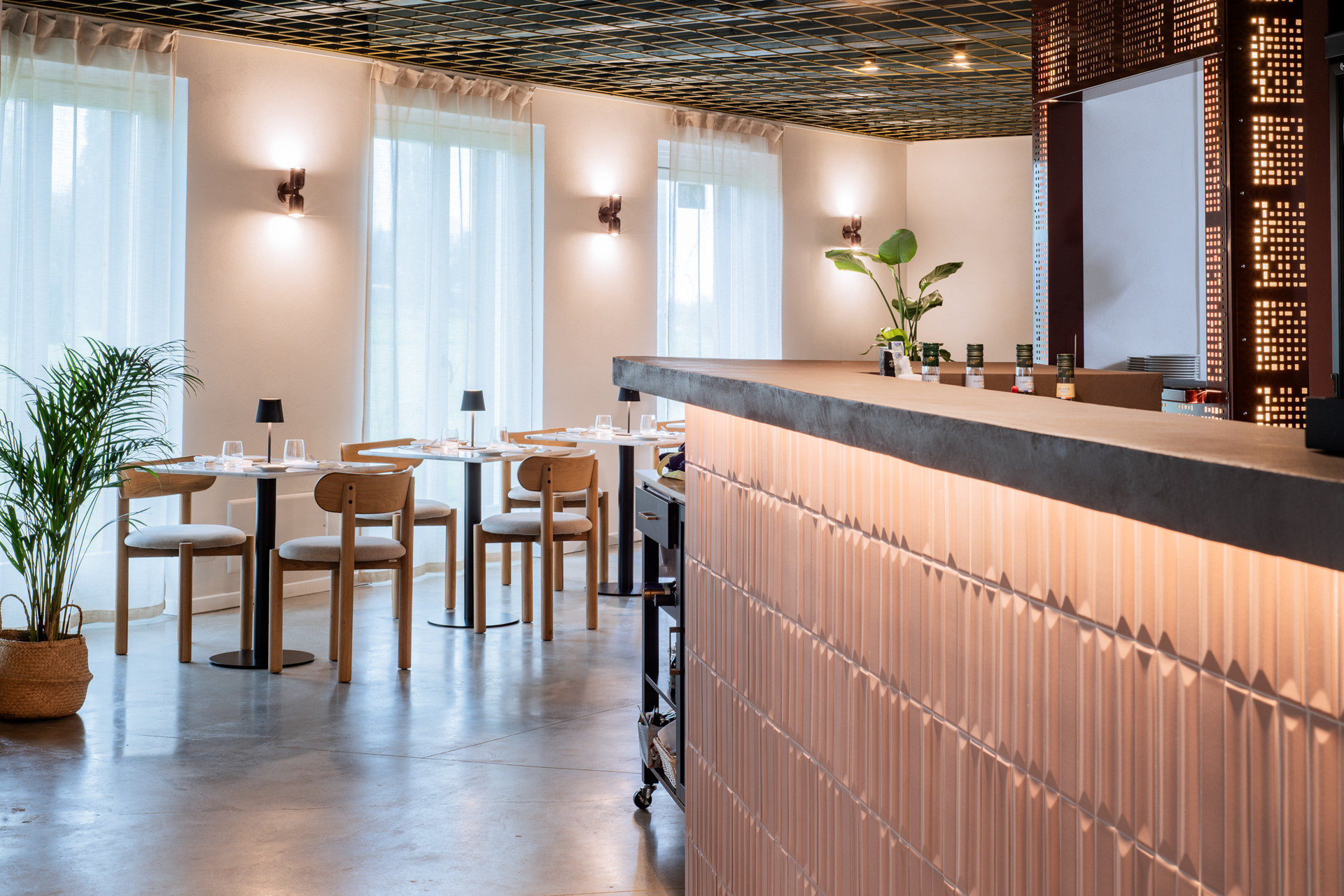 Interno del ristorante Archipelago con bancone bar rivestito con piastrelle 3d color terracotta con prospettiva verso le finestre. Soffitto con griglia ottonata a contrasto con il blu scuro fa da contrasto con le pareti e la griglia metallica color porpora a destra. Sulle pareti illuminazione Las Solas color porpora come la griglia del bancone.