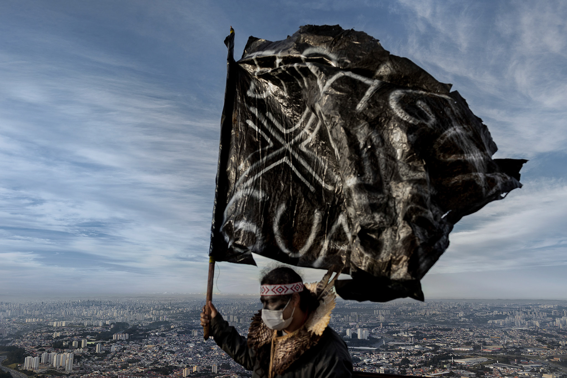 An unidentified Guarani Mbyá warrior raises a flag crafted from discarded plastic at the top of Pico do Jaraguá, the highest geographic point in São Paulo, Brazil, on June 30, 2021. This protest, featuring hundreds of indigenous ancestral inhabitants from surrounding villages, aimed to disrupt the signal of telephone and television communication antennas as a means to bring attention to the encroachment on indigenous territorial rights.
