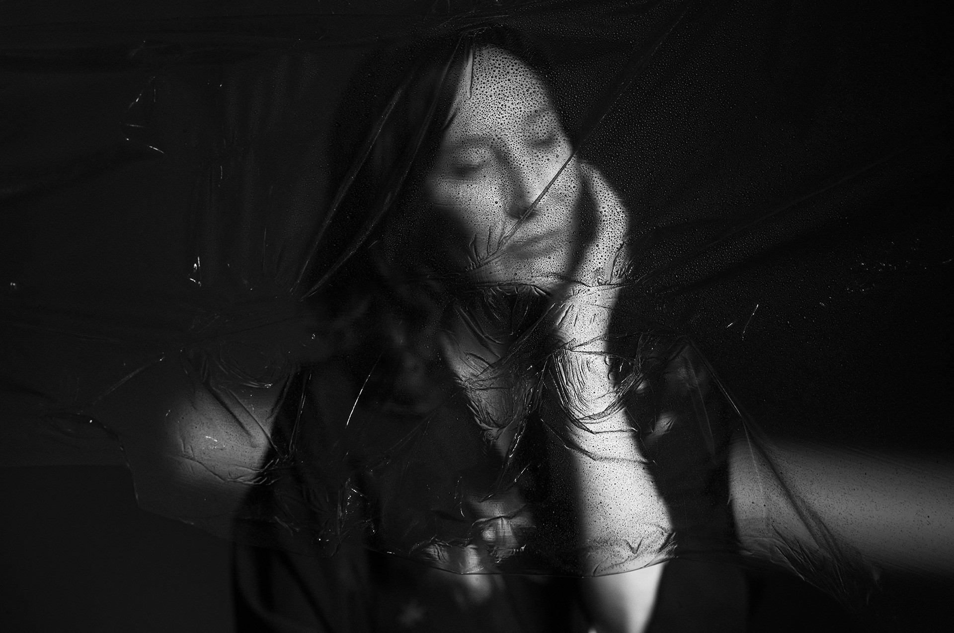 Portrait en noir et blanc : les yeux fermés, une femme appuie sa joue sur sa main derrière une matière transparente texturée.