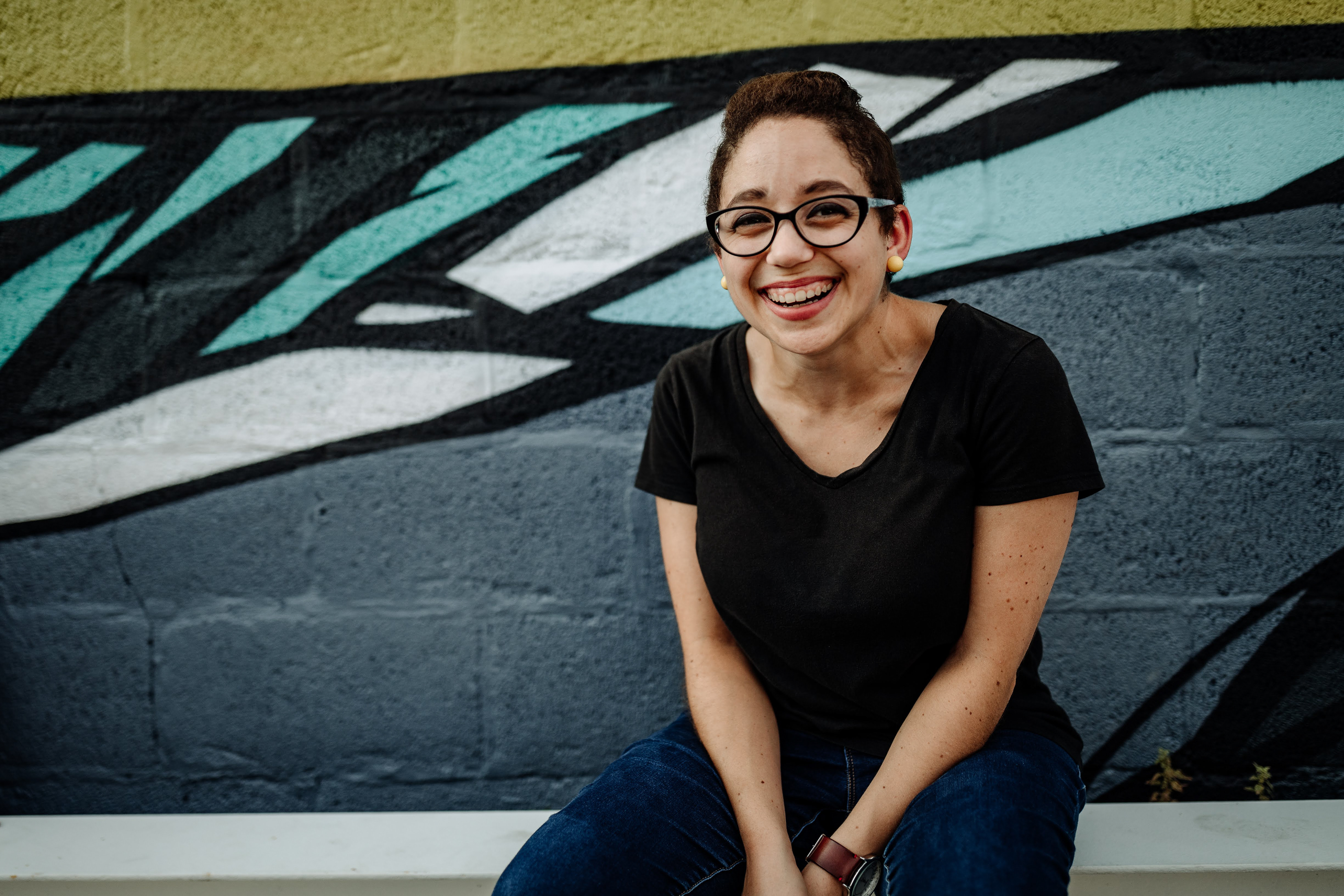 Becky, a bi-racial young woman with pixie-cut hair, a black shirt, and jeans, is seen smiling in front of a graphic-style mural.