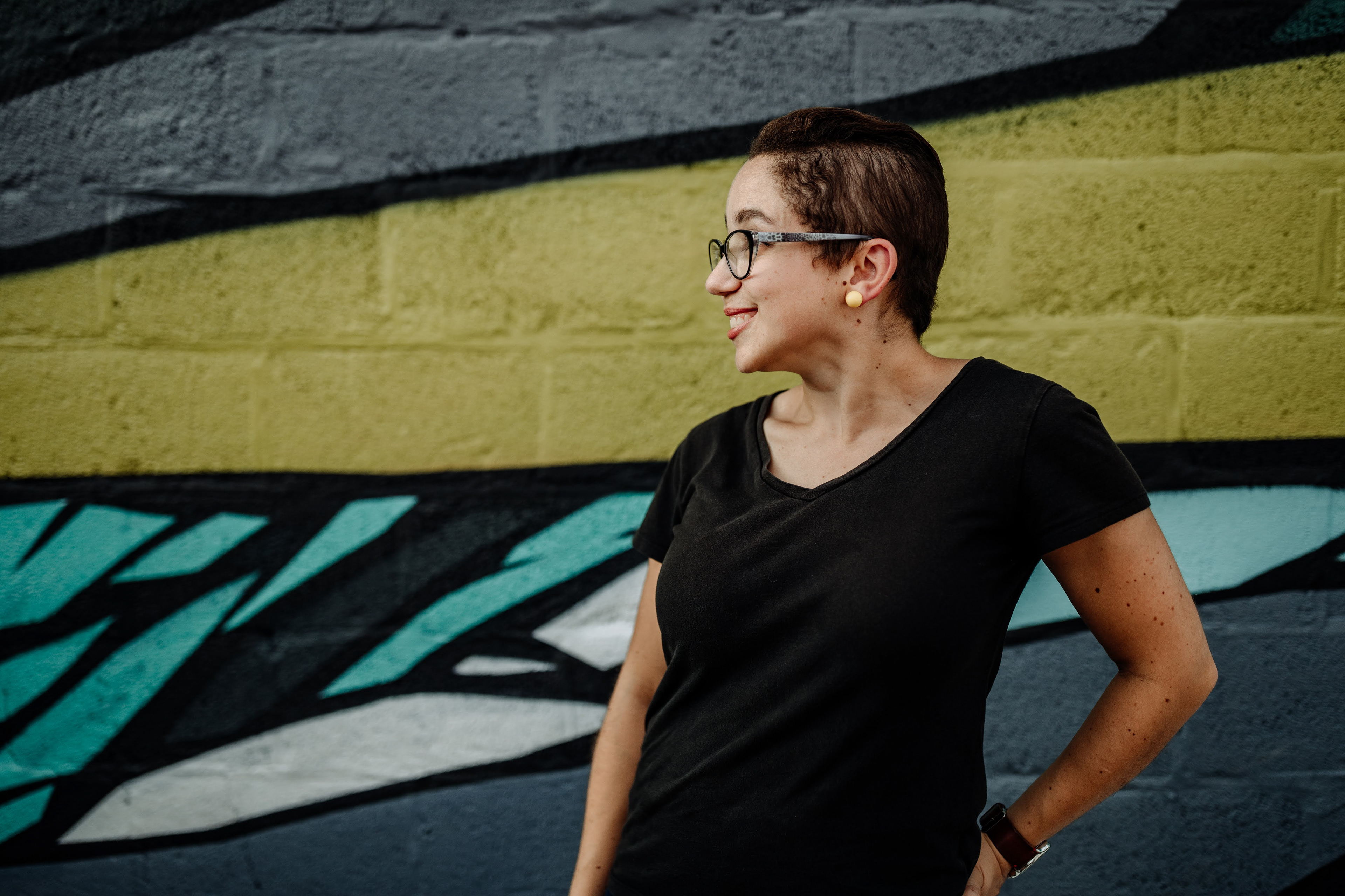 Becky, a bi-racial young woman with pixie-cut hair and black shirt, is seen looking to her right, smiling in front of a graphic-style mural.