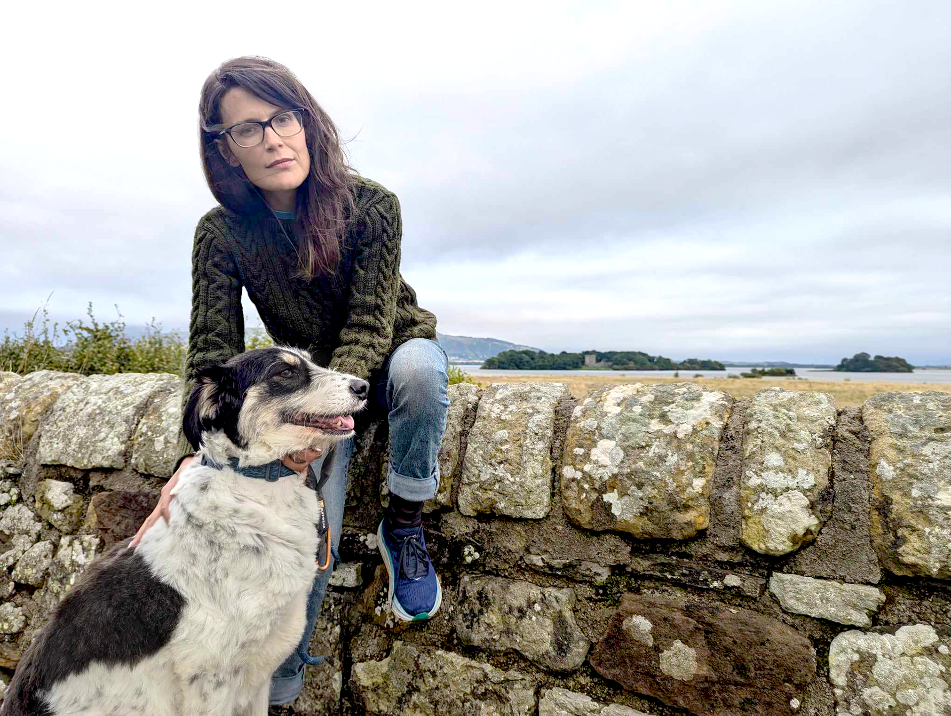 Morgan, a woman with long brown hair and glasses is sitting on a wall. She has her black and white dog sitting in front of her. In the back ground is Loch Leven Castle.