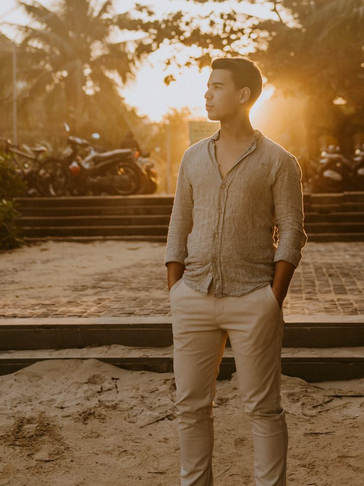 Photo of me standing on the sand at My Khe beach in Danang, Vietnam, with a warm sunset in the background. I am positioned in front of a few steps leading to the beach, with the street behind me creating an alley-like view. The sunset casts warm, golden light, illuminating the scene as I look towards the ocean, slightly turned to the left.