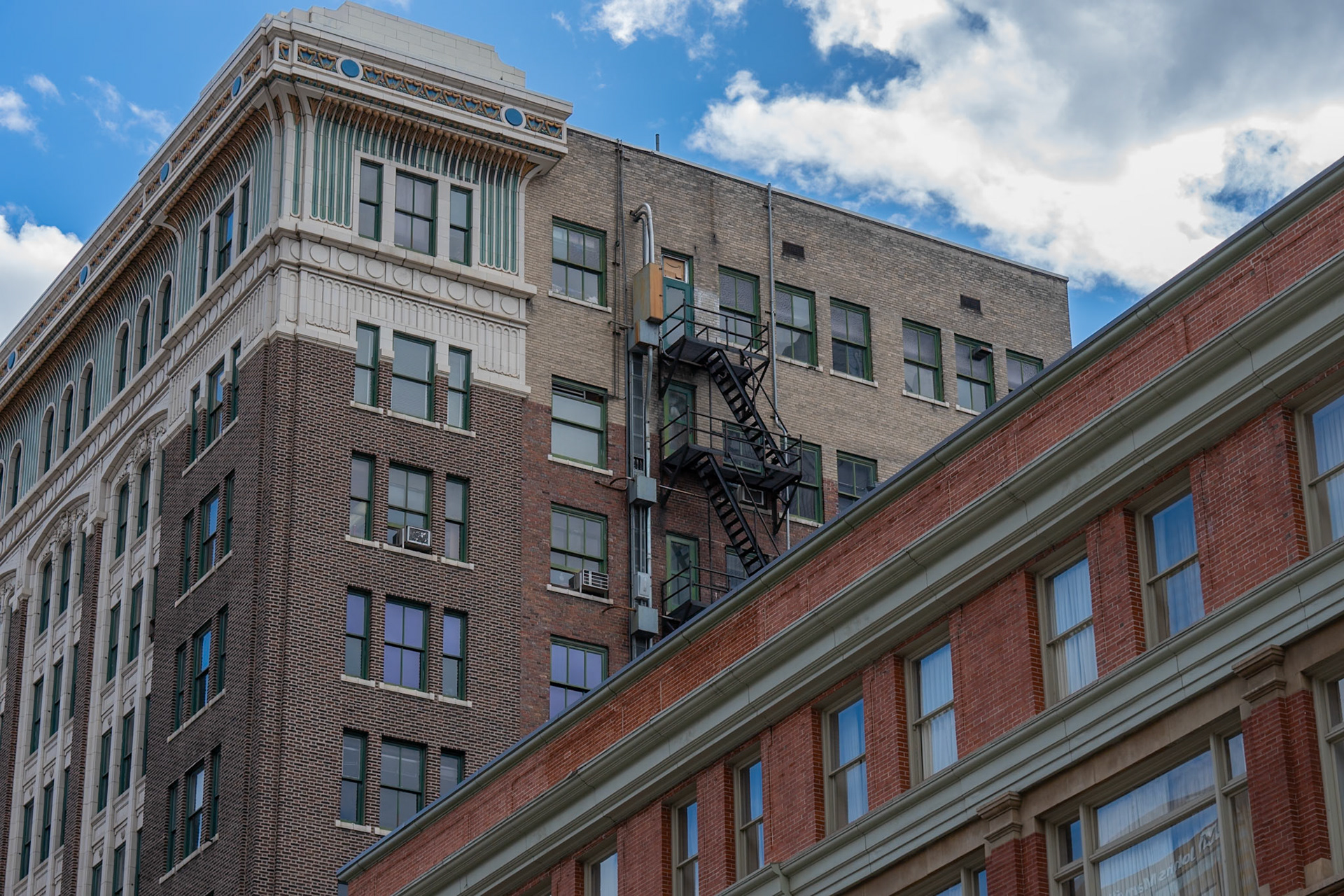 A view of urban architecture featuring classic brick buildings and a fire escape staircase against a partly cloudy sky, showcasing historic and vintage design elements.