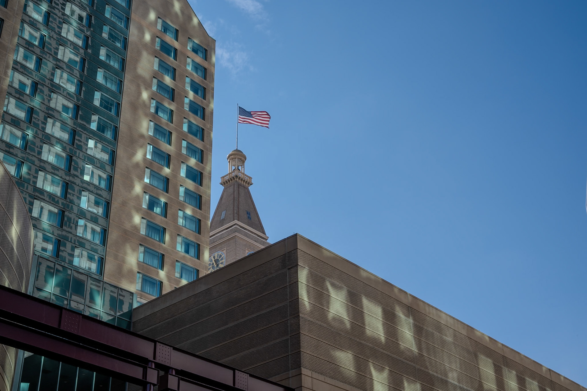 An urban scene featuring a modern building, with an American flag waving on top against a clear blue sky. The architecture showcases contemporary design elements and national symbolism.