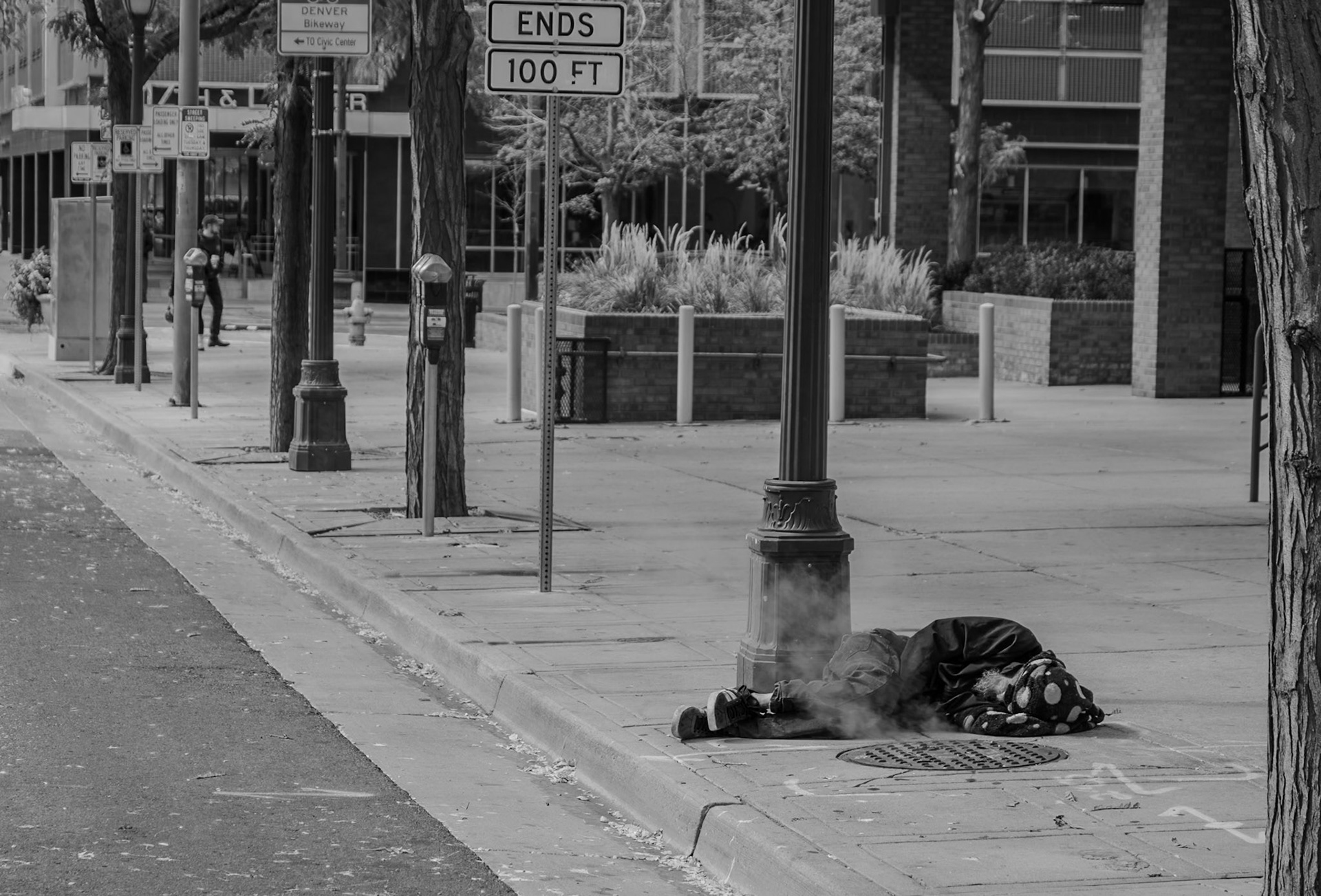 A black and white photograph depicting a quiet urban street scene with a person lying near a lamppost. The atmosphere suggests solitude and urban life.