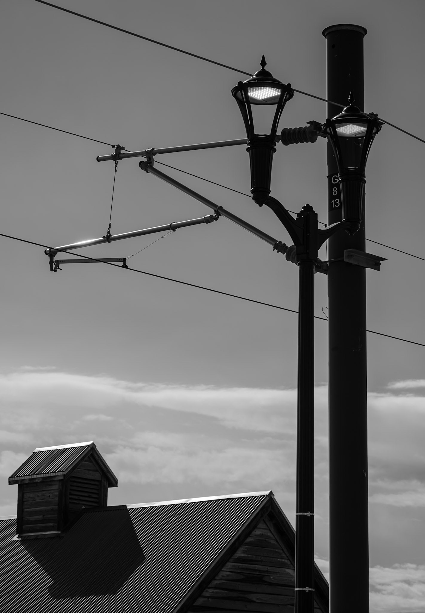 Monochrome image showcasing an antique street lamp against a clear sky, with part of a wooden building and power lines visible, evoking a nostalgic and historic feel.