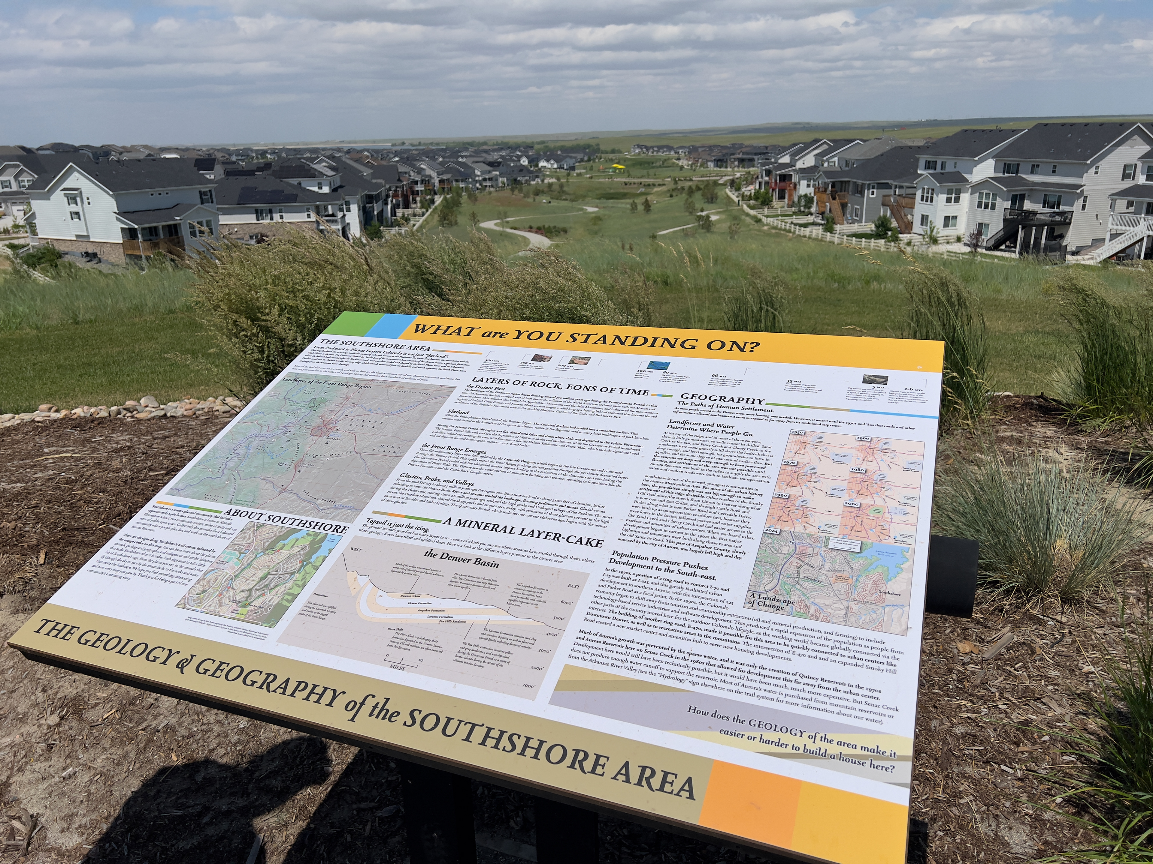 A large interpretive sign showing it in front of a large suburban housing development with parkland. The sign's topics covers geology and geography of the area.