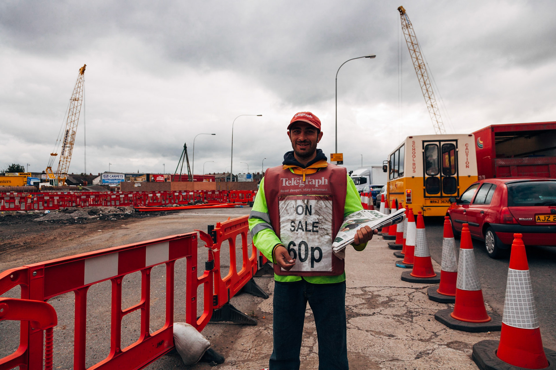 The Belfast City Tour. Un immigrato di origine asiatica vende giornali lungo la strada in prossimità di nuovi quartieri in costruzione.