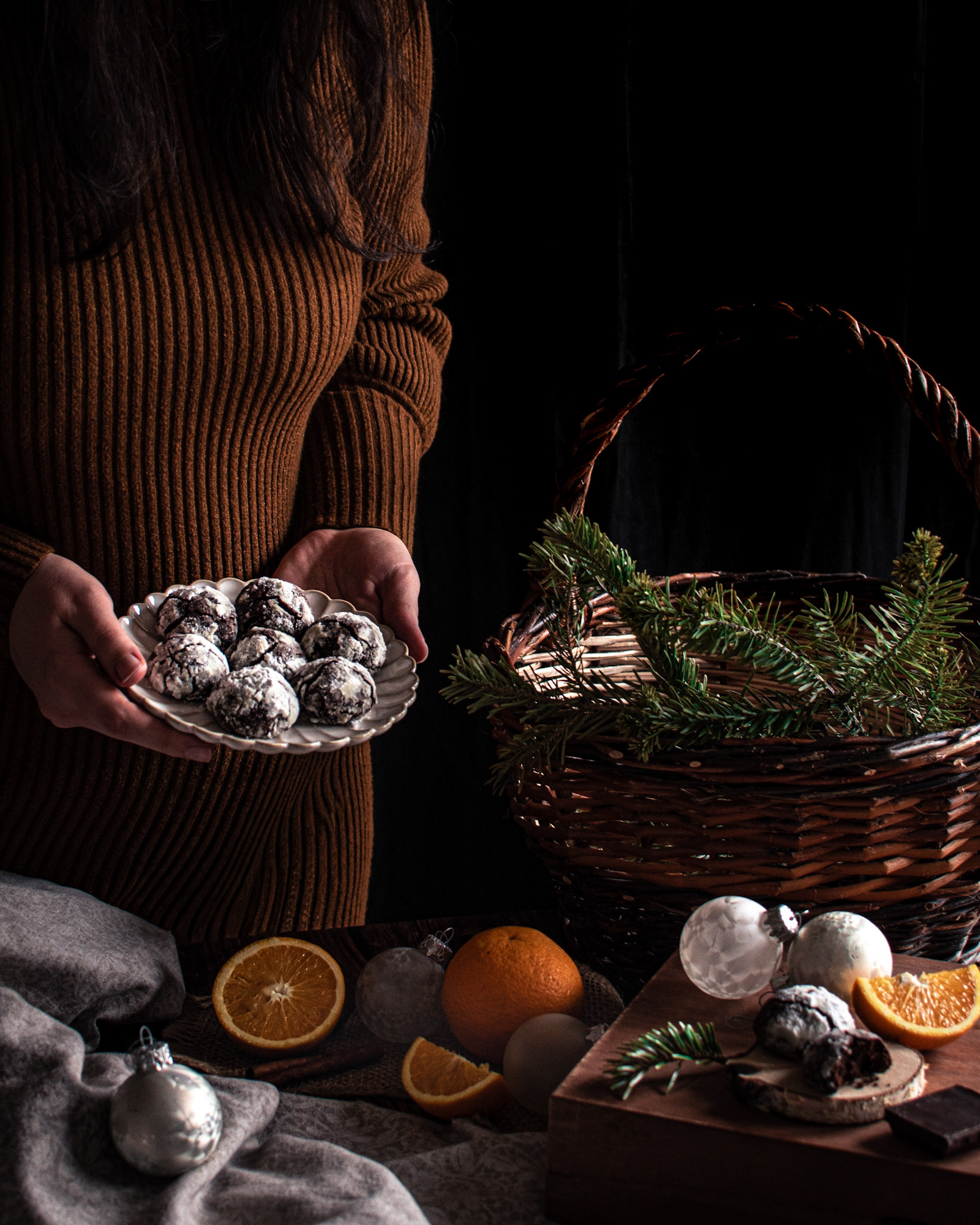 a woman in brown dress holding a plate with crinkle cookies, basket with pine branches, oranges, Christmas ornaments