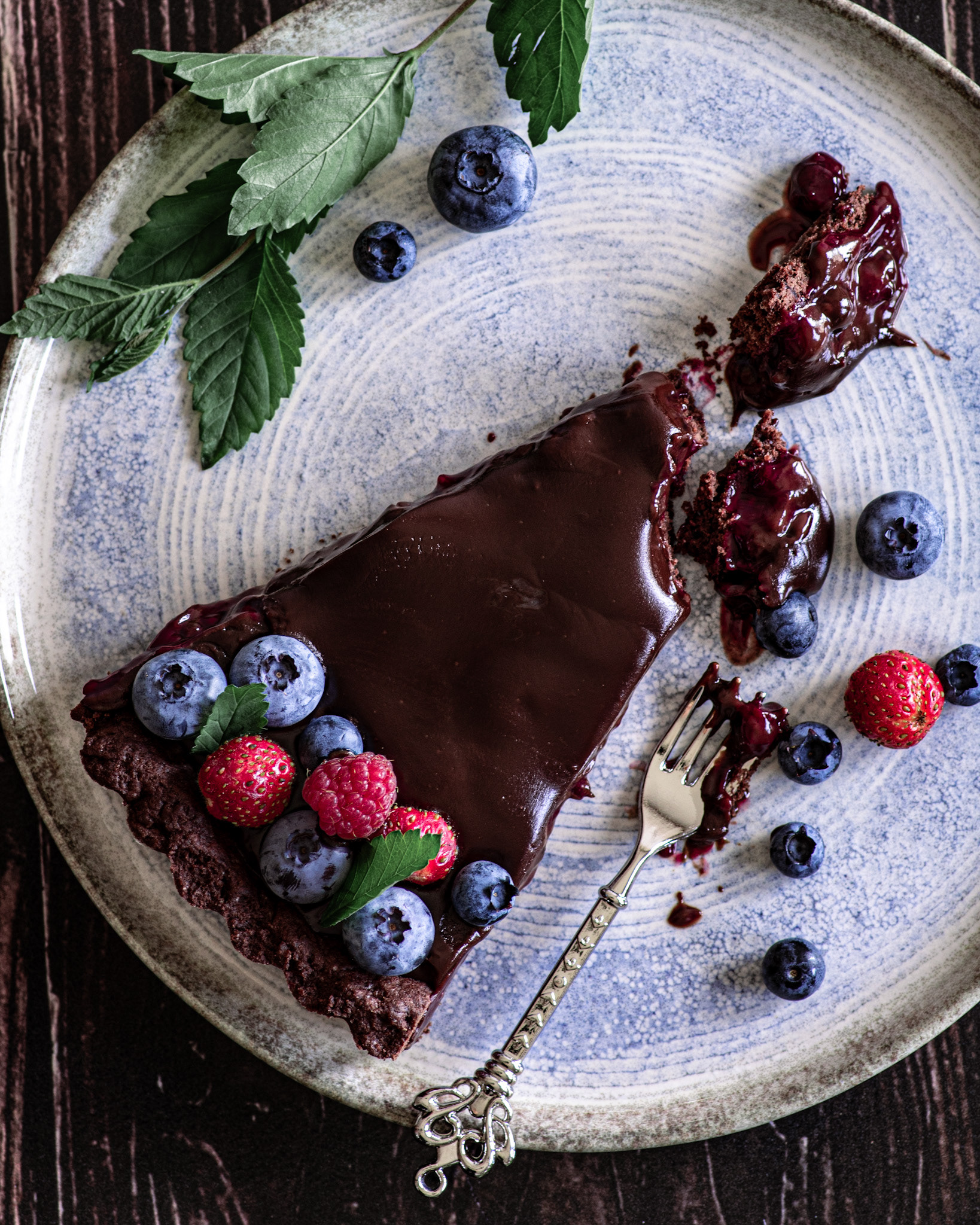 chocolate tart with berries, a messy slice, decorated small forks, blue plate and greenery