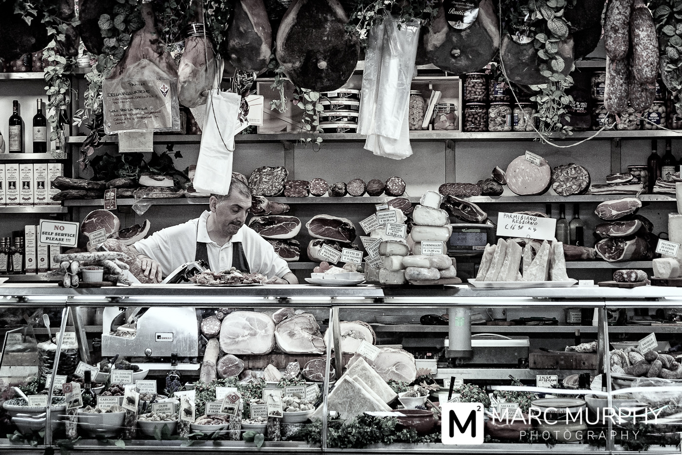 Italian Businessman Slicing Meat Behind the Counter in Florence Italy with Hanging Meat, Fresh Cheeses and Herbs
