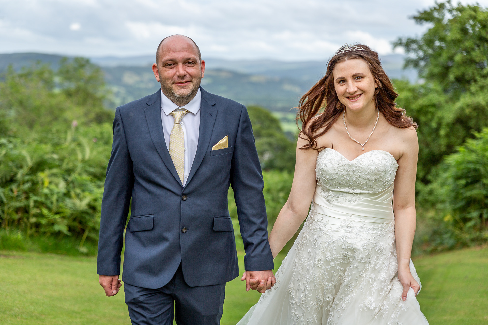 A bride and groom walking holding hands across Bala Golf Club on their wedding day. The bride is wearing a strapless white wedding dress and the groom is wearing a dark blue suit and yellow tie.