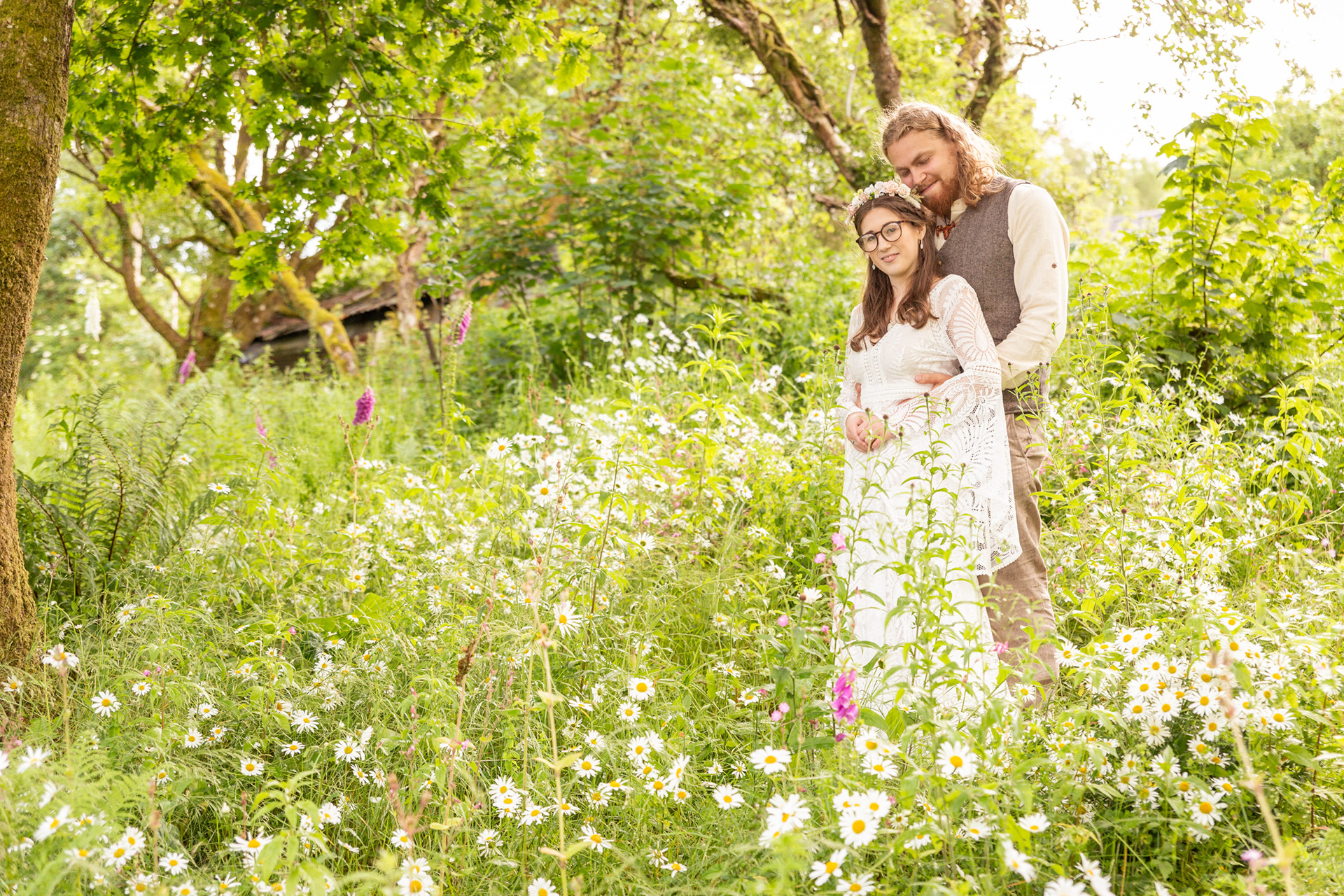 Portrait of a bride and groom during a woodland wedding in Dolgellau. Bride is in a white wedding dress. The groom is standing behind his bride with his arms around her waist. They are standing amongst wild flowers in a woodland.